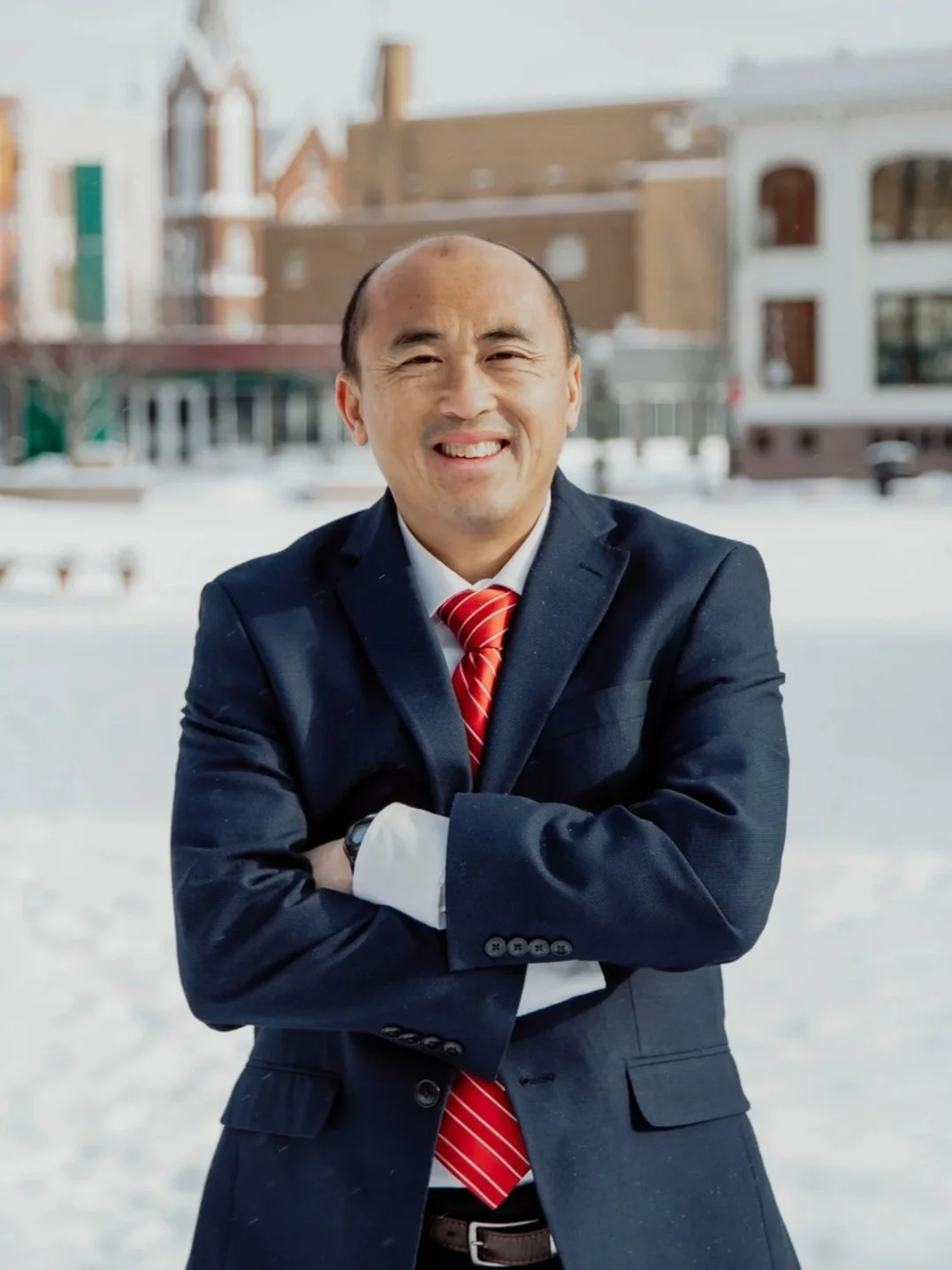 A man in a business suit with a red striped tie standing outdoors in a snowy area, smiling with arms crossed.
