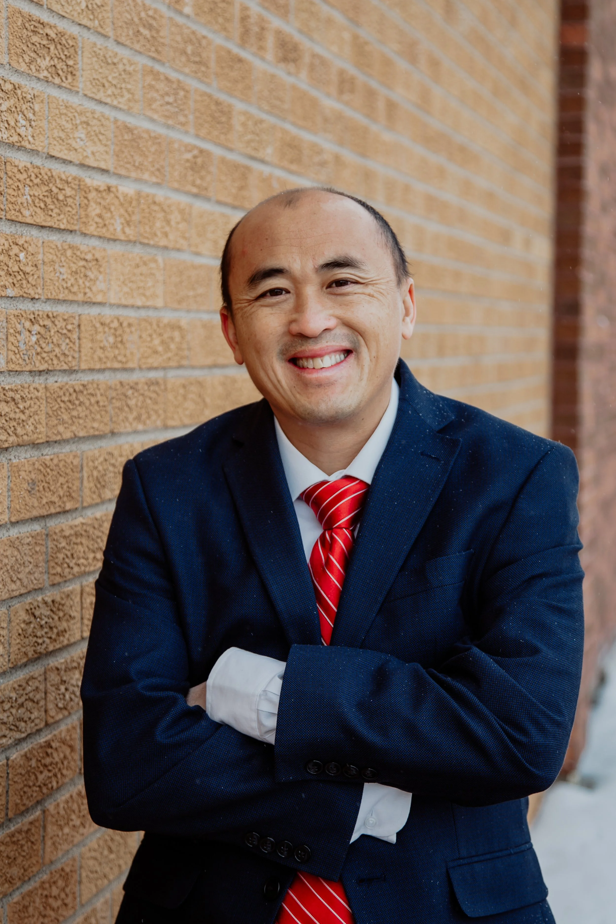 A smiling man in a dark business suit with a red and white striped tie, standing against a brick wall with his arms crossed.