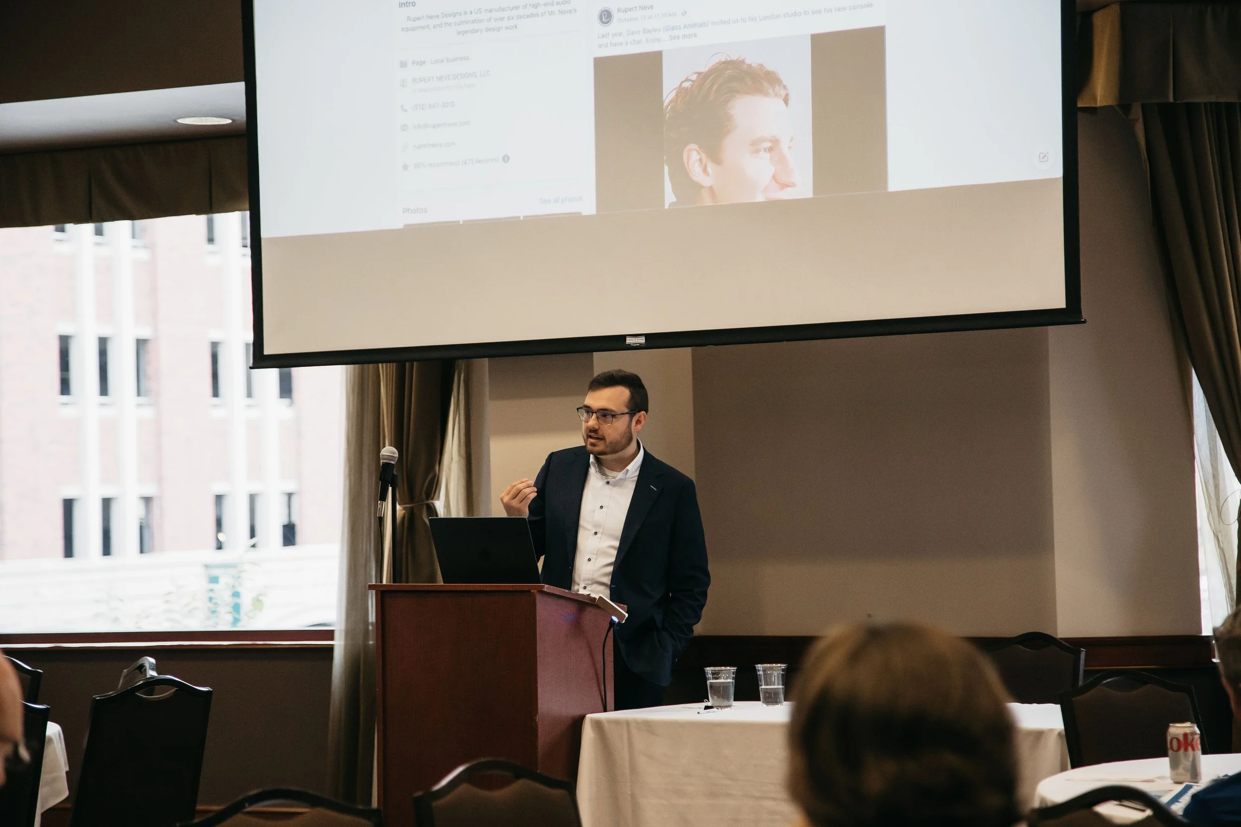 A man in a suit and glasses is giving a presentation at a podium with a microphone in a conference room. A large screen shows a slide with a profile picture of a person's head and website information. The room has large windows with buildings visible outside, and tables with glasses of water and a soda can are in foreground.
