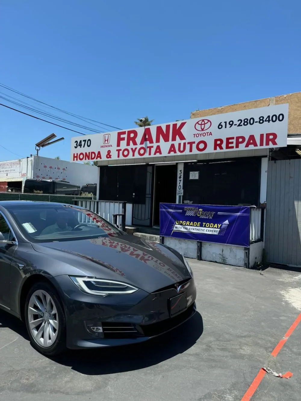 Exterior view of Frank Honda & Toyota Auto Repair shop with a black Tesla car parked in front, blue sky overhead, and signage displaying the business name and contact information.