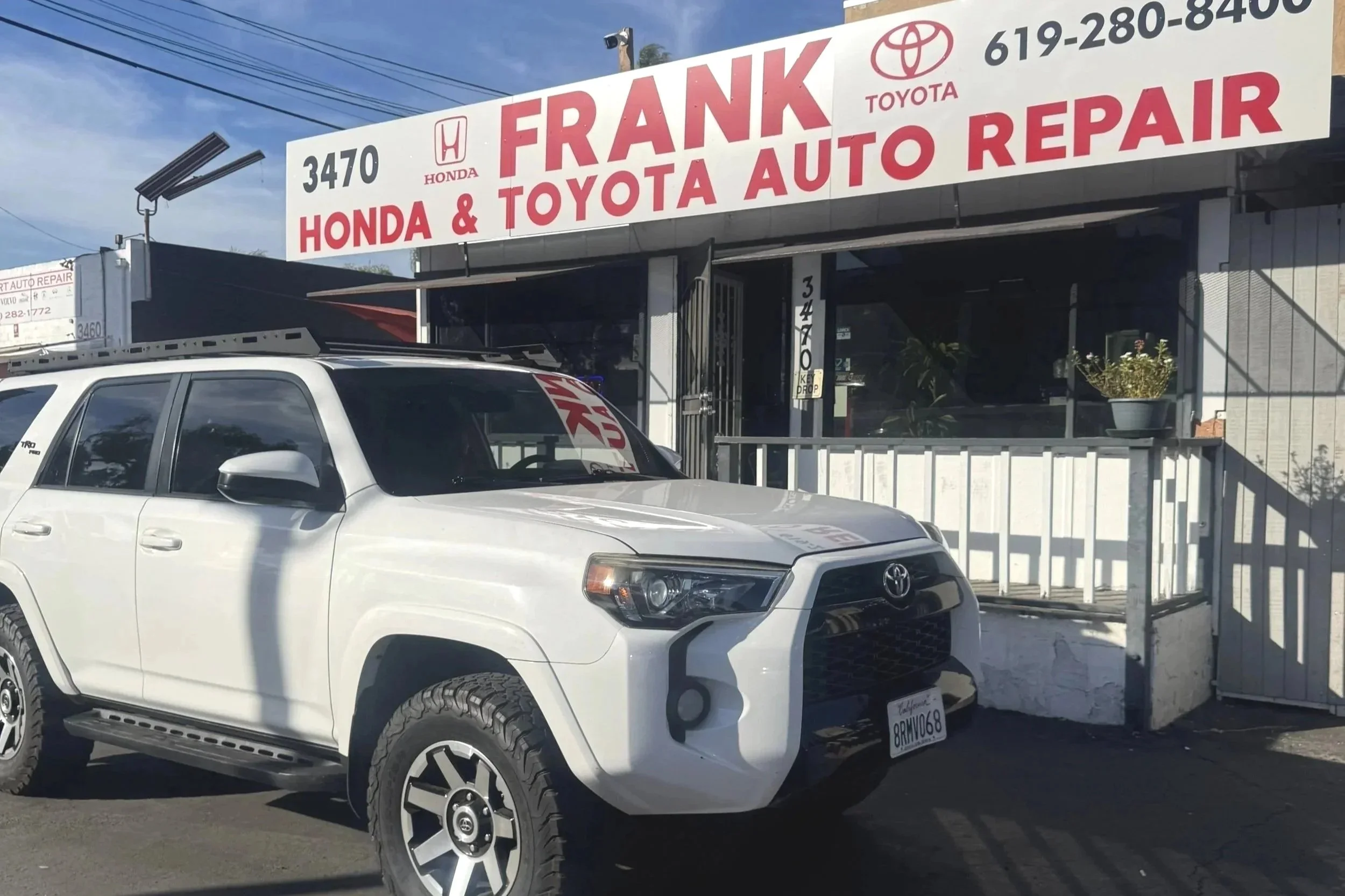 A white Toyota SUV parked outside a sushi restaurant with a red and white sign that reads "Frank Honda & Toyota Auto Repair." The restaurant has a white exterior with potted plants near the entrance and a driveway in front.
