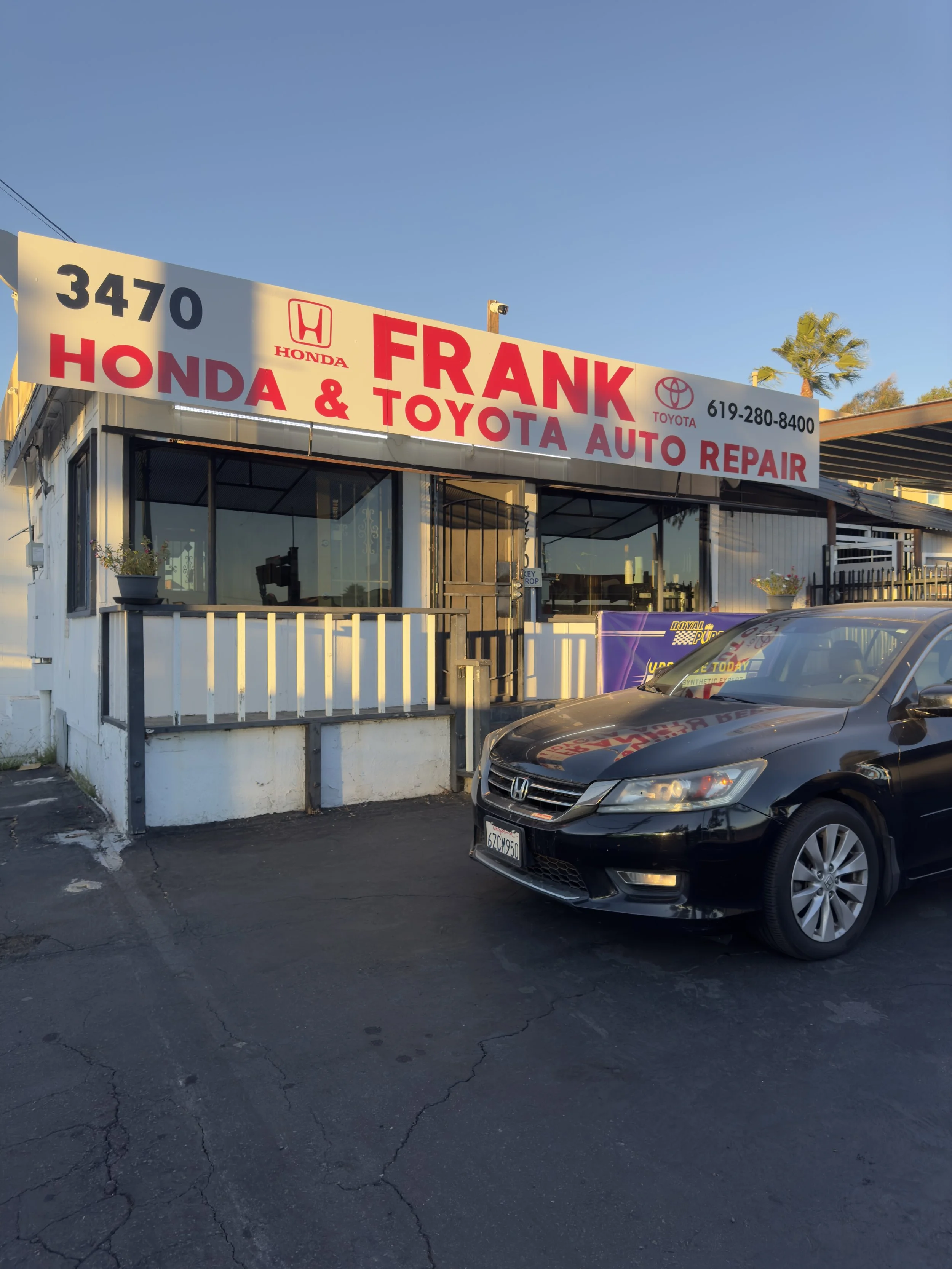Front view of a small auto repair shop named 'Frank Honda & Toyota Auto Repair' with a black Honda car parked outside, sunlight casting shadows, palm trees in the background.