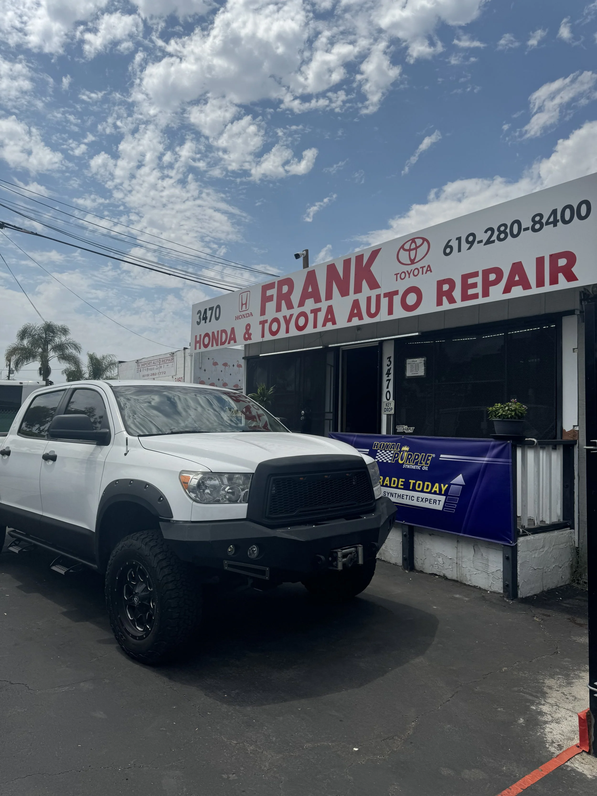 A white pickup truck parked outside Frank Honda & Toyota auto repair shop with a sign displaying the shop's name, contact number, and advertisements for synthetic oils and car trade services, under a partly cloudy sky.