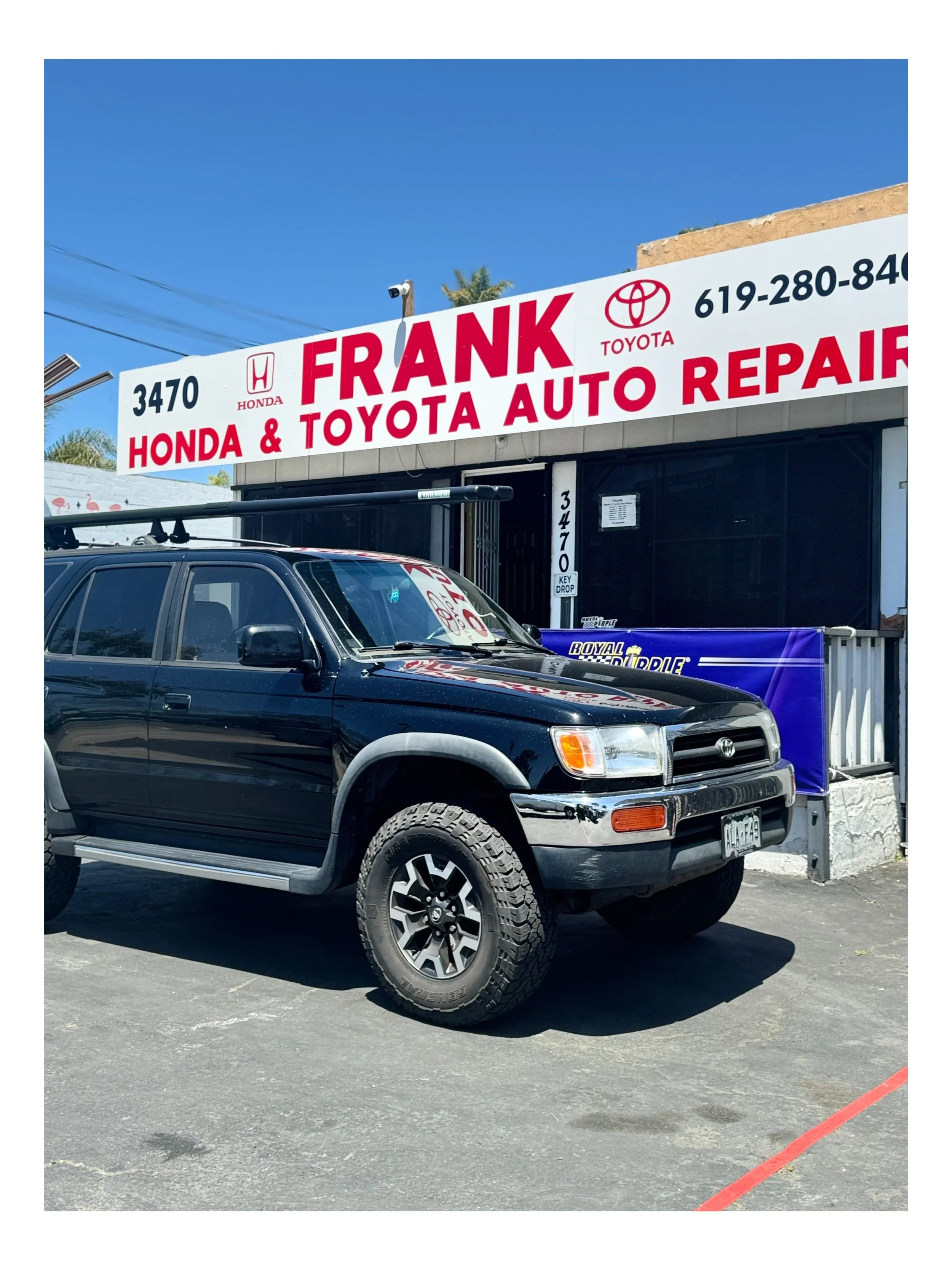 A black Toyota SUV parked outside Frank Honda & Toyota Auto Repair shop with a large white sign overhead displaying the shop's name and contact information.