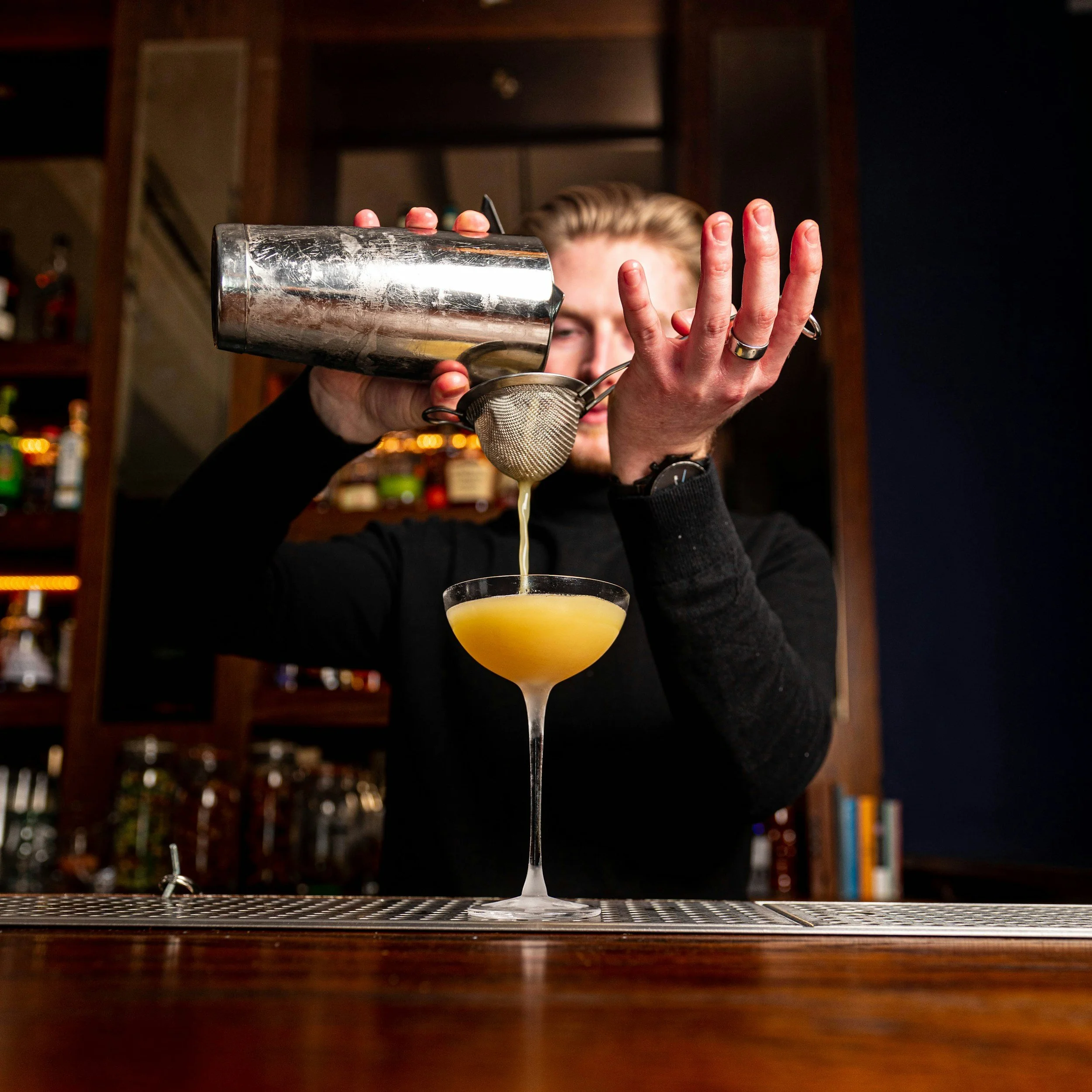 A bartender pouring a yellow cocktail through a strainer into a coupe glass at a bar.