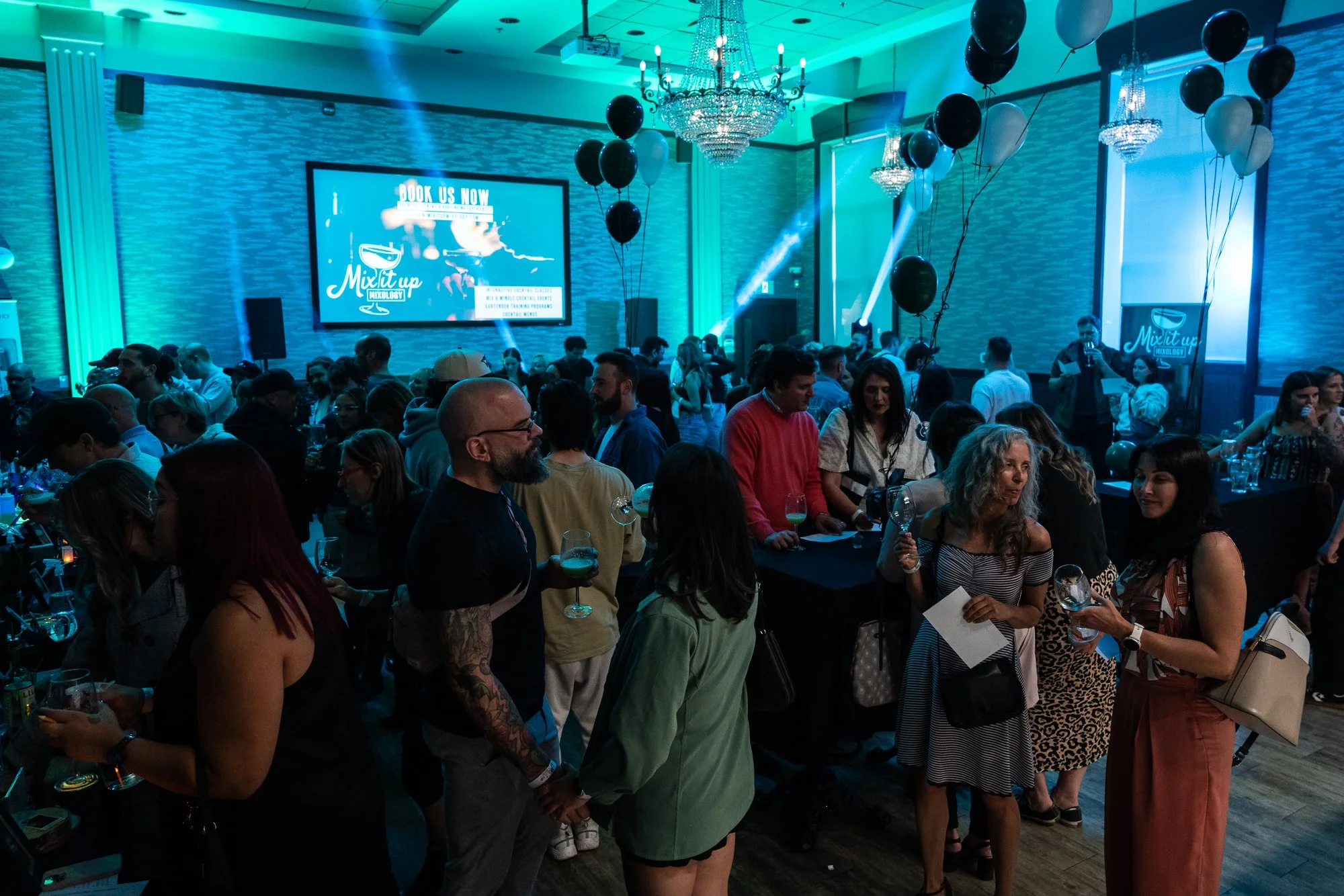 Crowd of people at a social event or party in a decorated venue with balloons, chandeliers, and large screen displaying promotional content.