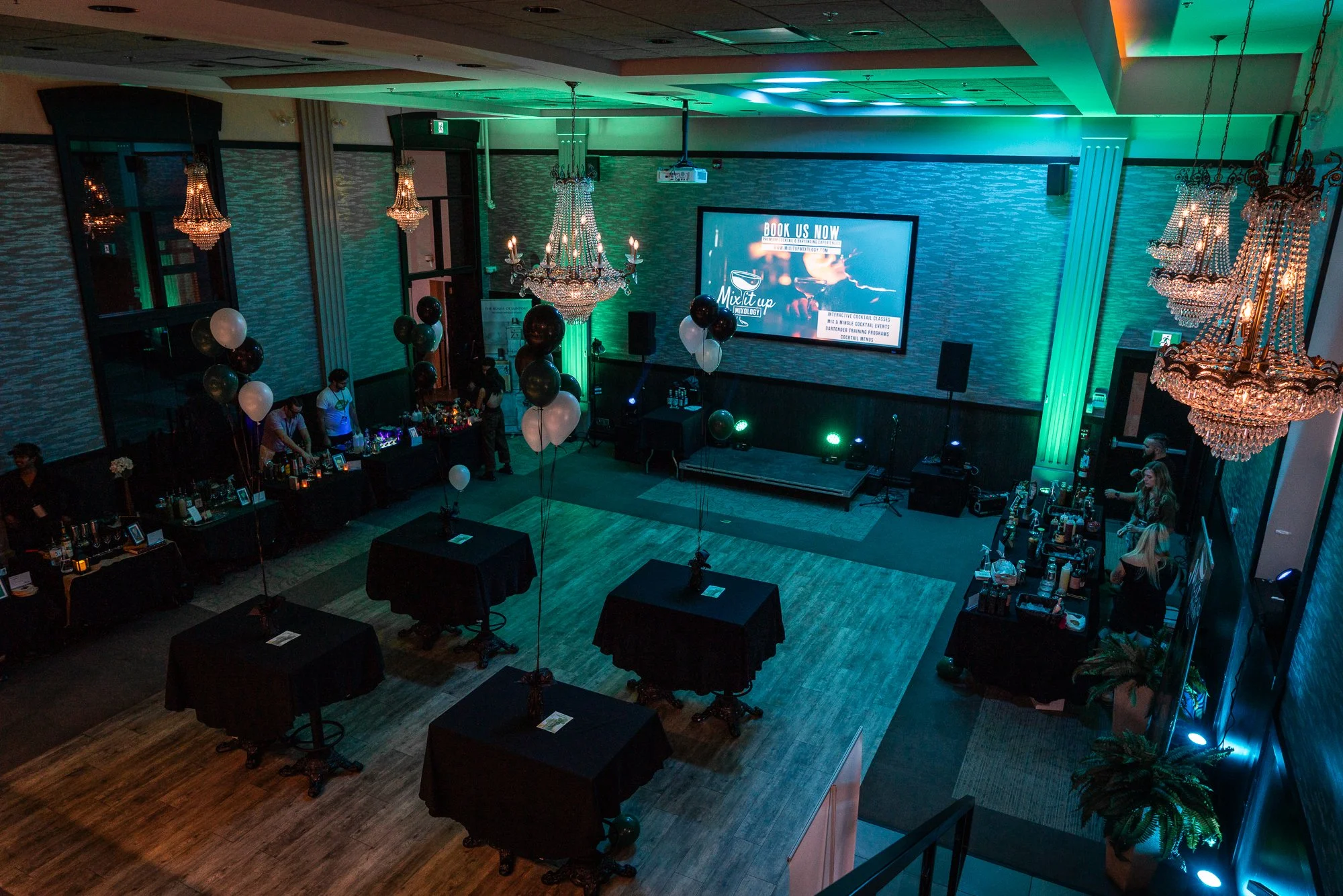 An event space decorated with chandeliers, black and white balloons, and tables with cocktail stations, set up for a party or celebration with dim lighting and teal accent lights.