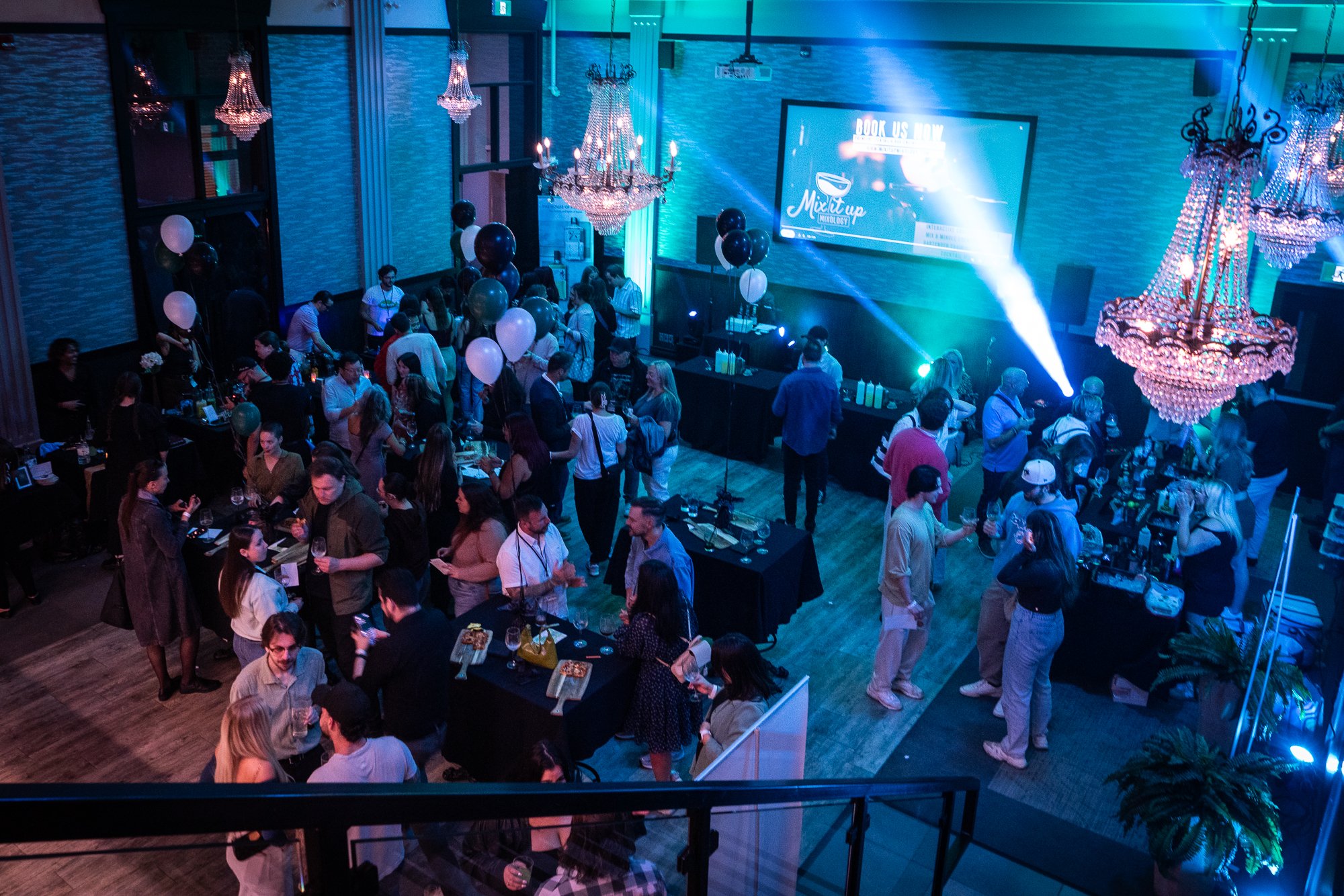 A lively indoor party scene with people socializing, balloons, chandeliers, and stage lighting in a large event hall.