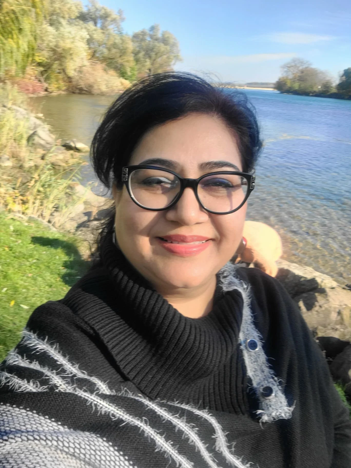A woman with glasses smiling outdoors near a river with trees and grass in the background.