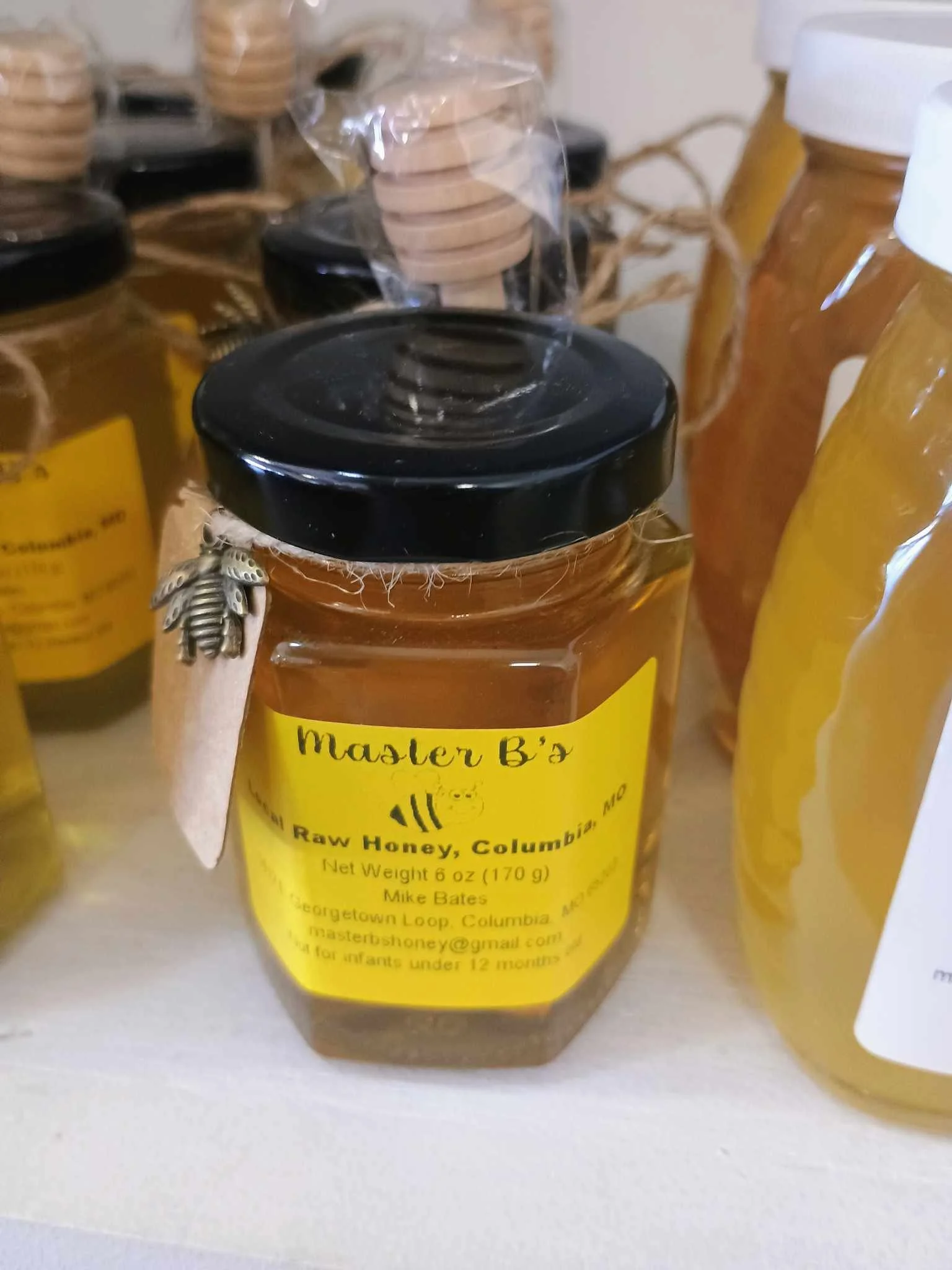 Jar of Master B's raw honey with black lid and yellow label, surrounded by other honey jars on a shelf.