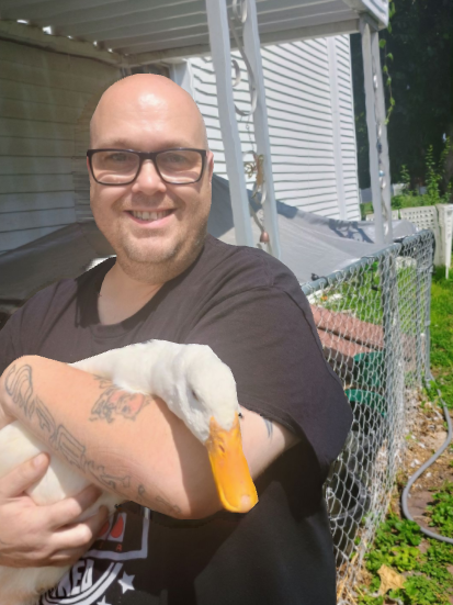 Man with glasses smiling and holding a white duck outside next to a house and a fence.