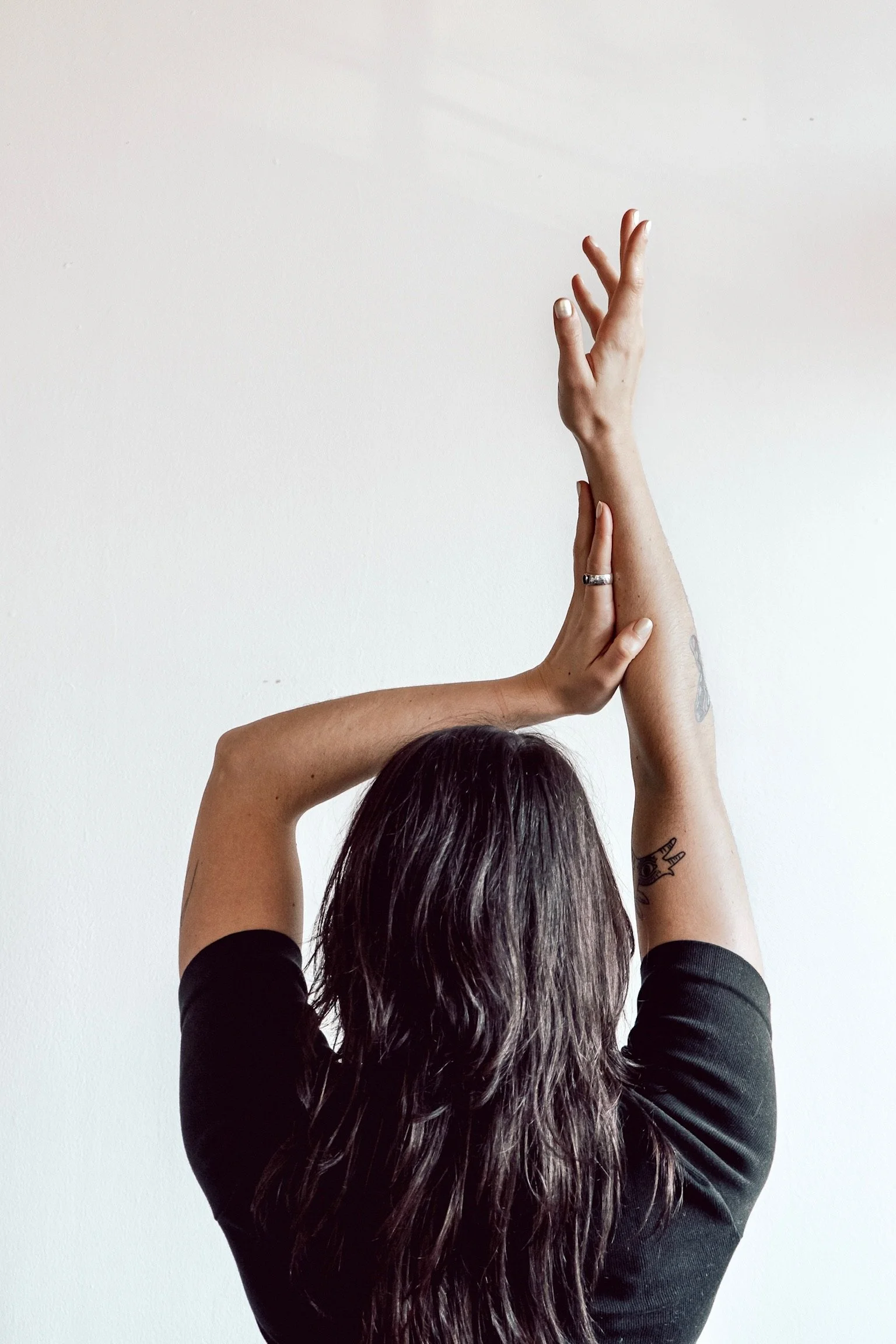 A person with long dark hair wearing a black shirt, raising both arms upwards with hands intertwined, standing in front of a plain white wall.