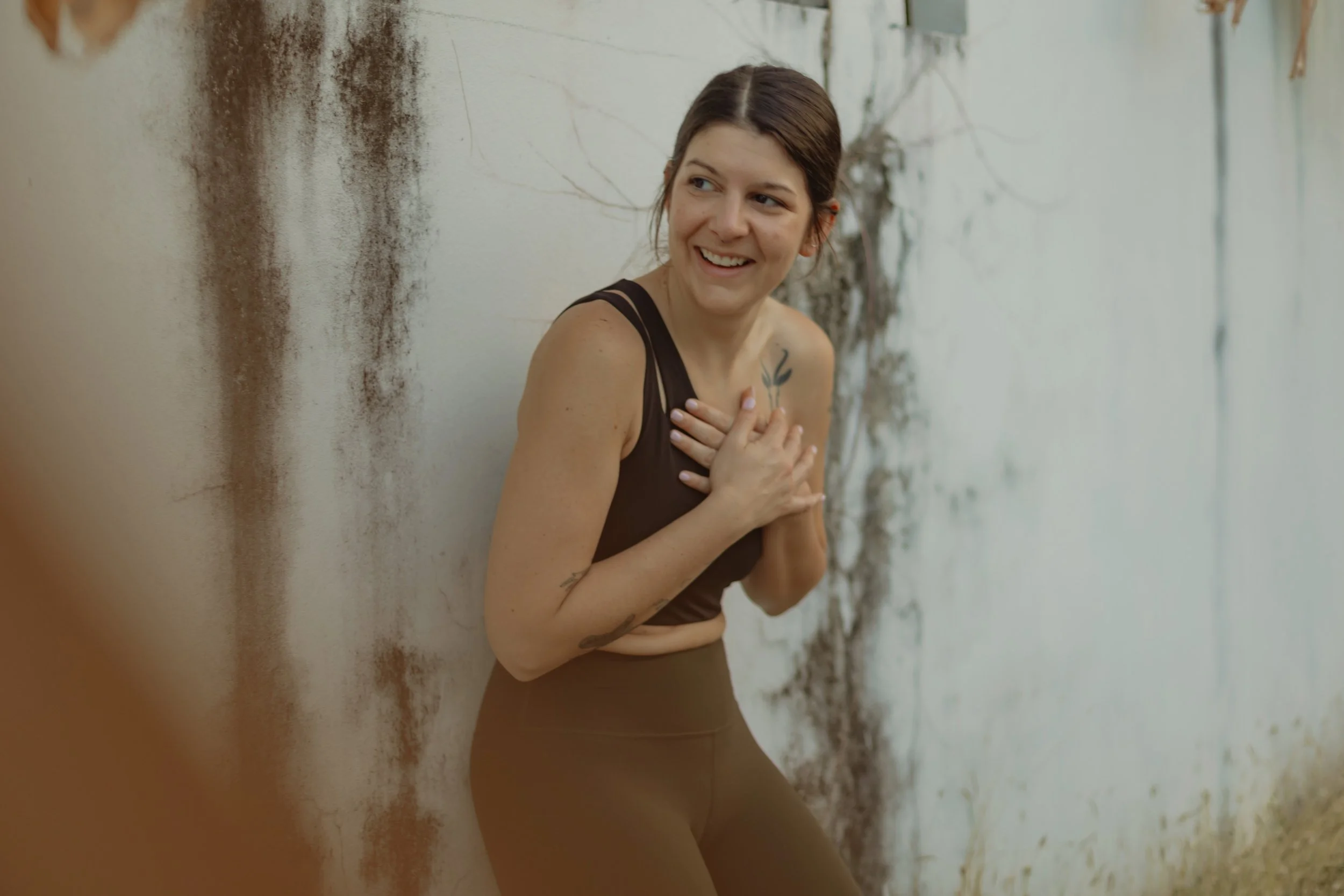 A woman with short brown hair, wearing a sleeveless black top, is smiling and holding her chest with one hand while leaning against a weathered white wall with peeling paint and some vines.