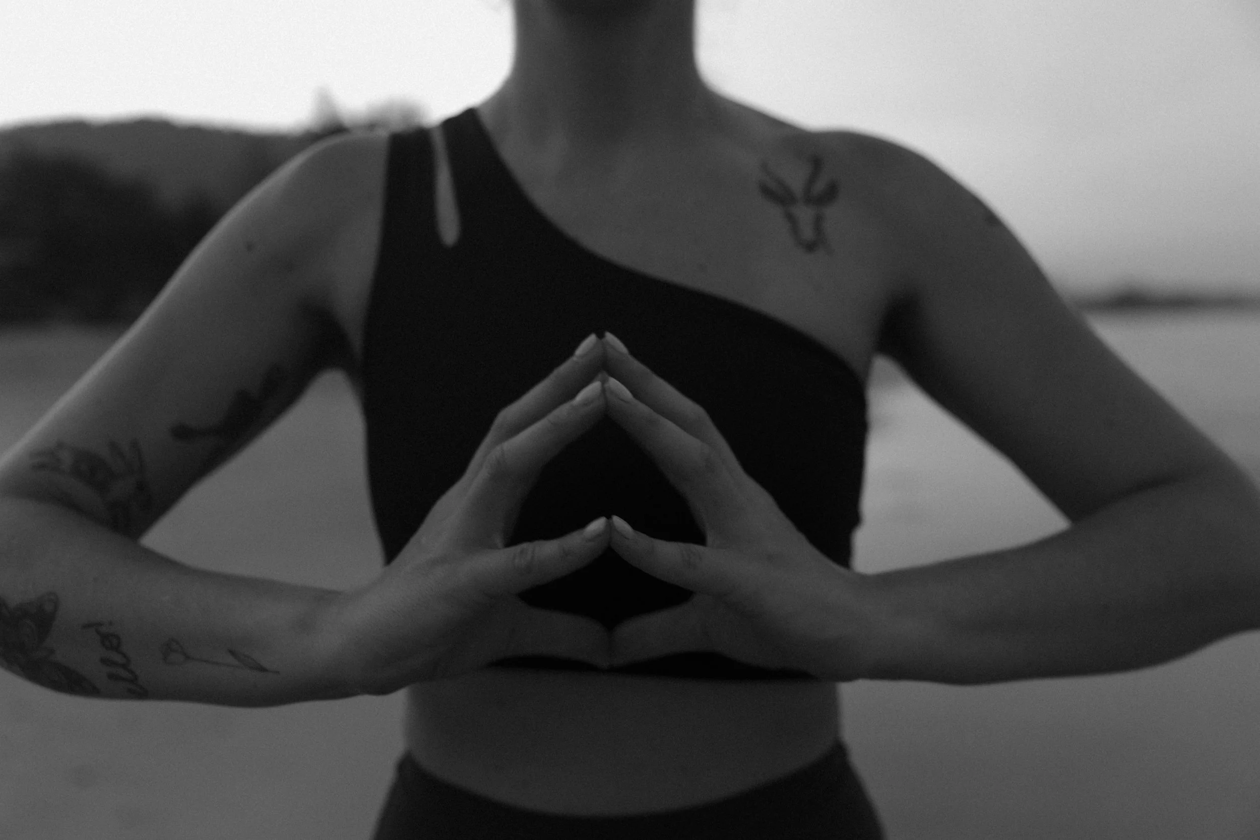 A person standing outdoors near water, with hands in the hakini mudra position and fingertips touching, forming a globe shape. The person is wearing a black sports top and has tattoos on their arms and shoulder.