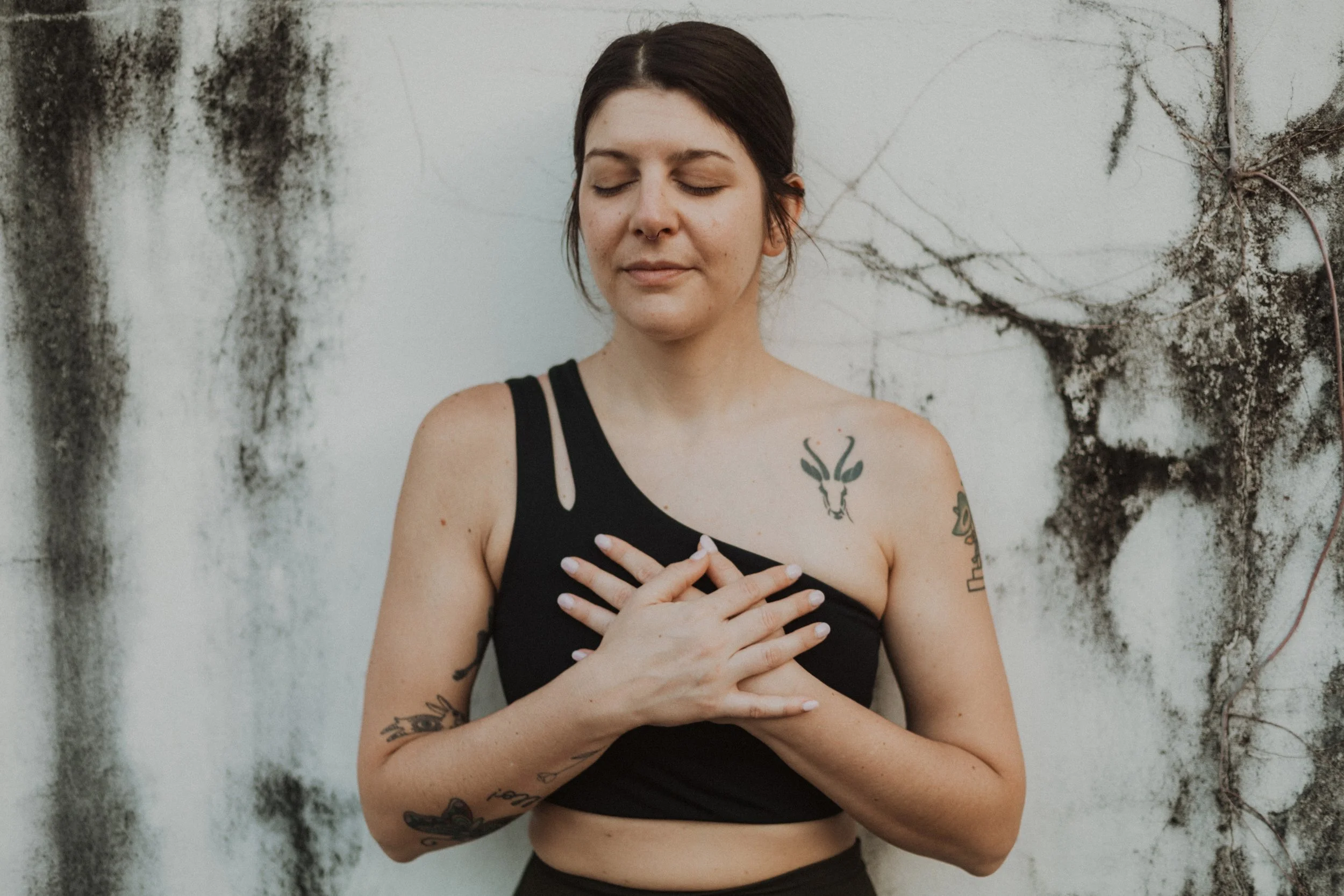 A woman with short dark hair, tattoos on her arms and chest, wearing a black tank top, standing with eyes closed and hands crossed over her chest against a weathered white wall with black mold and vines.