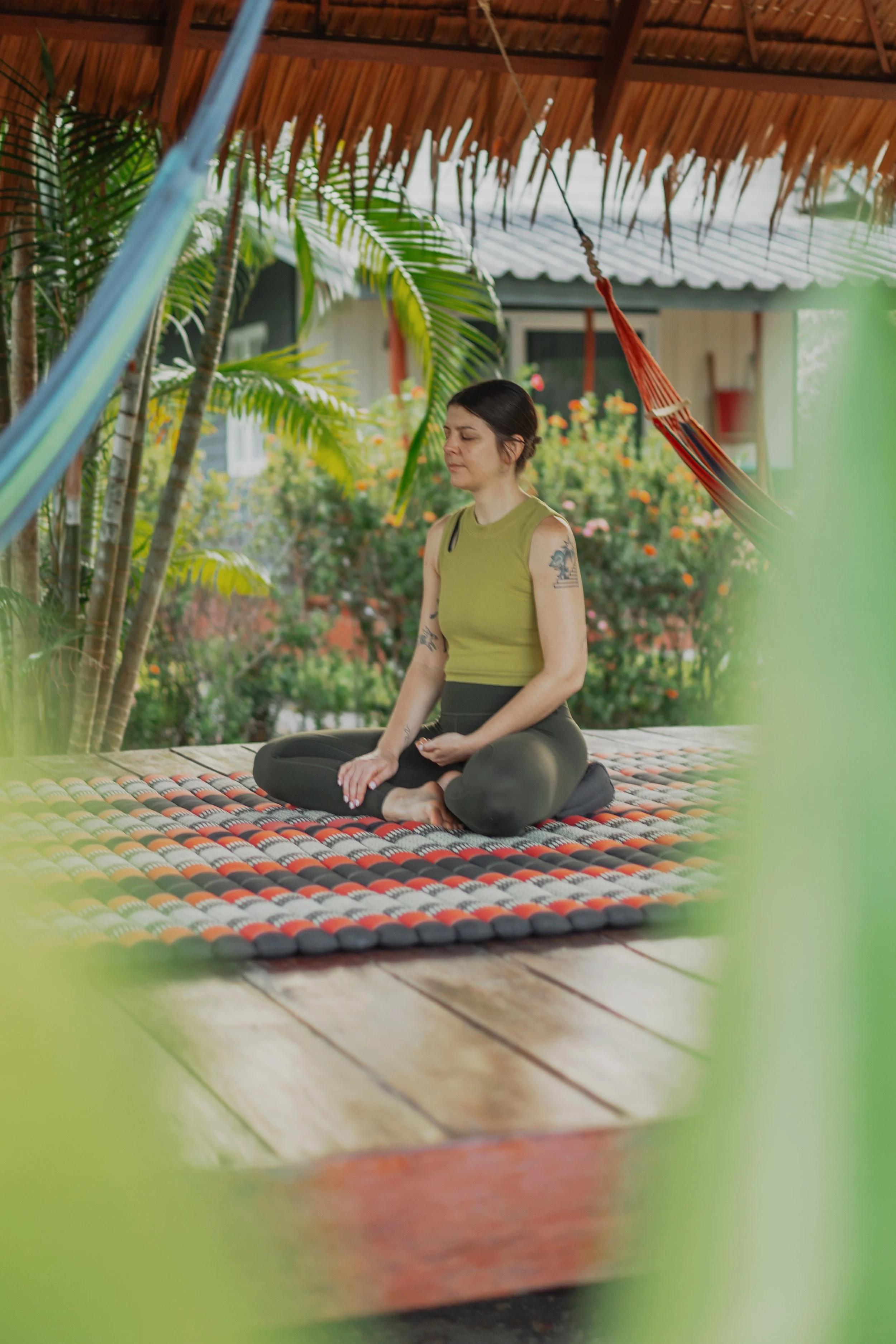 Woman practicing meditation outdoors on a colorful woven mat, surrounded by tropical plants and a porch with a hammock hanging from the ceiling, in a garden setting.