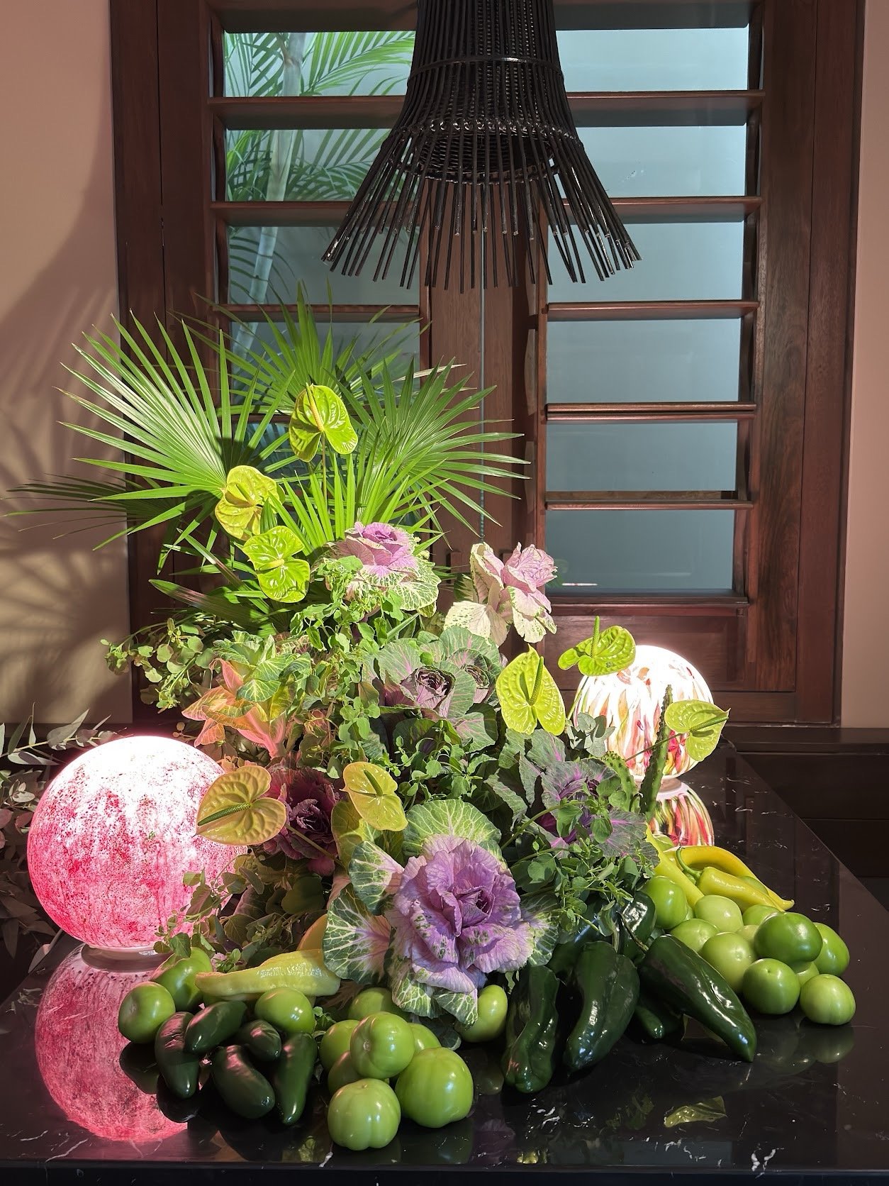 Indoor display of lush green plants, decorative pink and white lamps, and fresh vegetables including green apples, avocados, and peppers against a dark surface with a wooden framed window in the background.