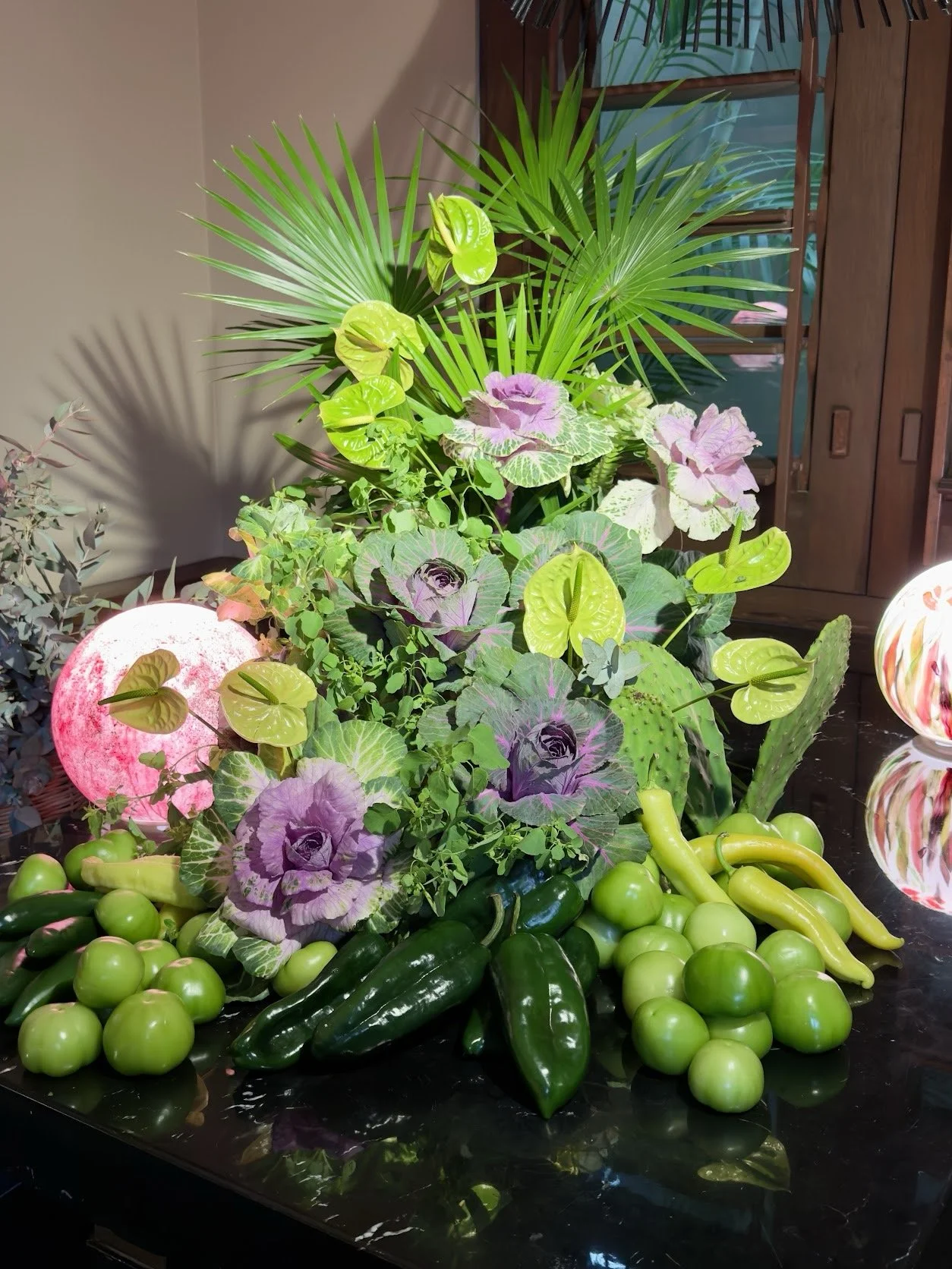 Arrangement of lush green plants with purple and green ornamental cabbages, green chili peppers, cucumbers, green tomatoes, and a glowing pink moon-shaped light. A wooden cabinet and a shutter are visible in the background.