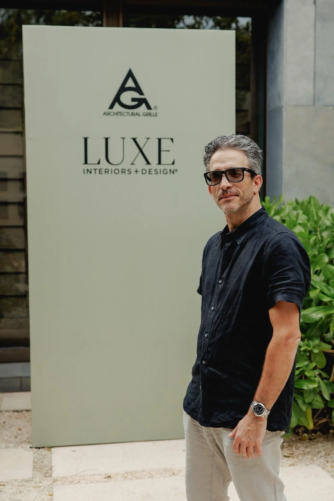 Man wearing a black shirt, beige pants, black sunglasses, and a watch, standing outdoors in front of a large sign that reads "LUXE INTERIORS + DESIGN" at a Donna Perillo Design event in Mexico.
