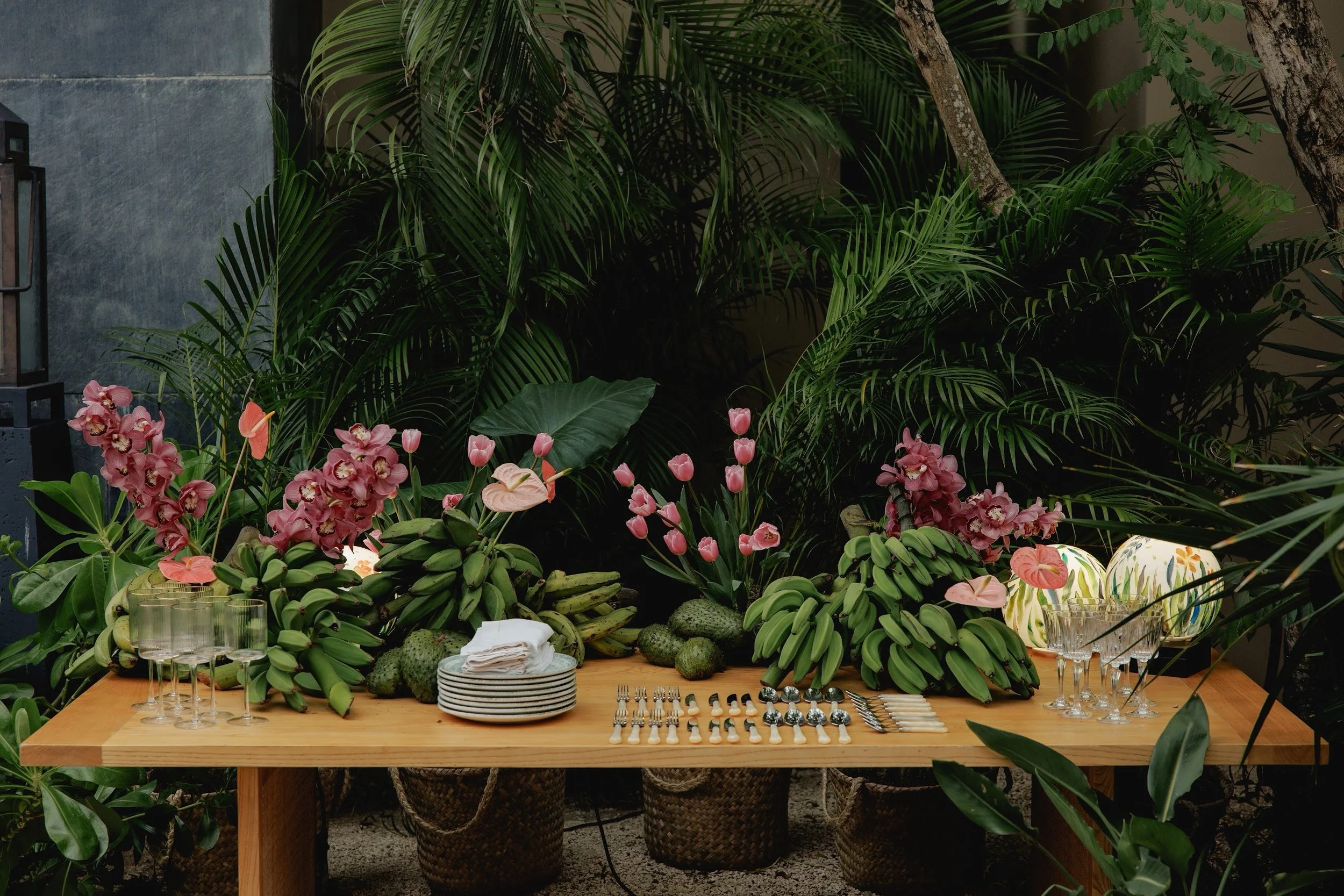 A table set for a party or gathering with plates, glasses, and cutlery, crowded with green bananas, pink orchids, and various plants and foliage in the background.