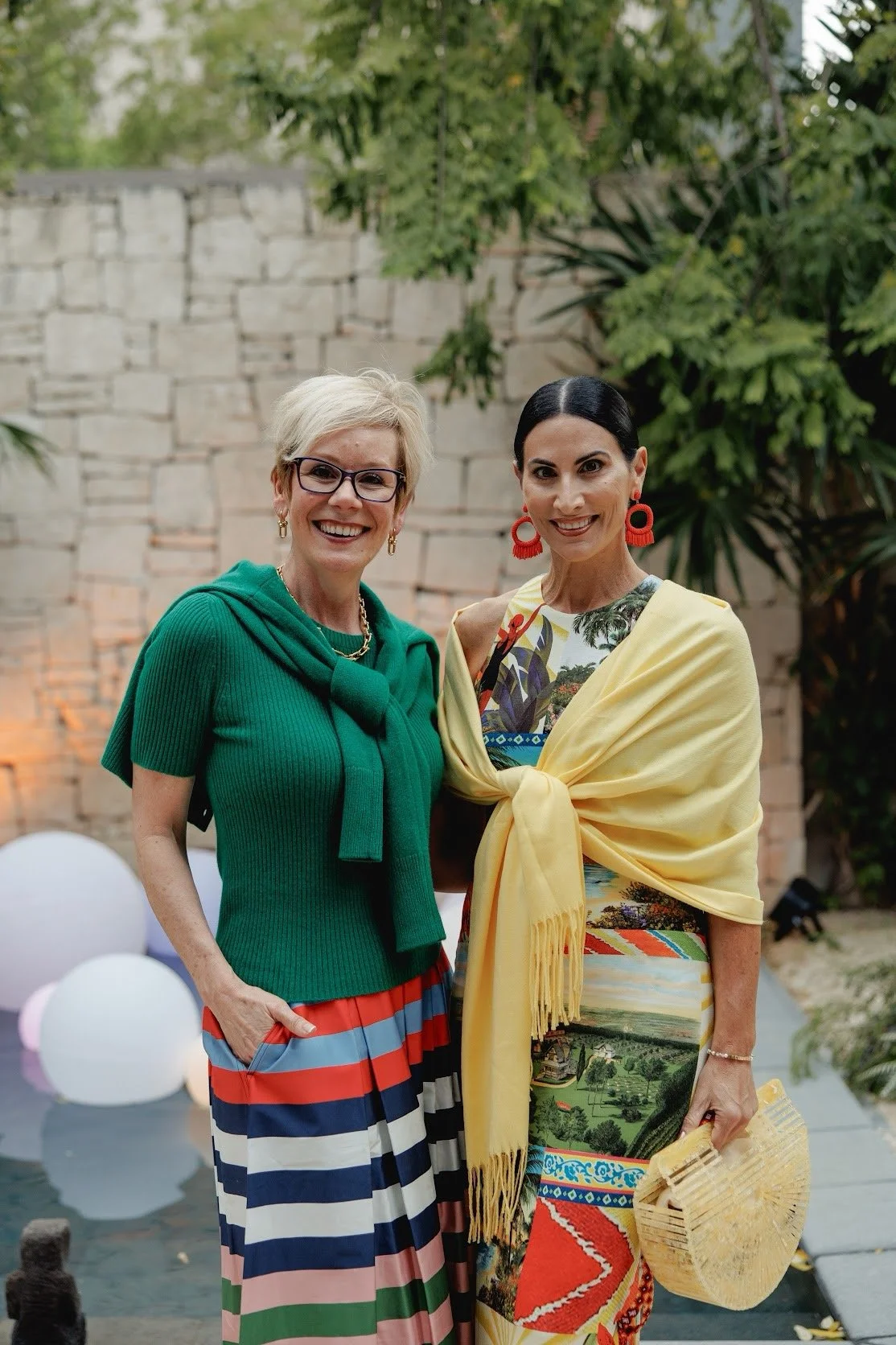 Two women smiling outdoors with trees and a stone wall in the background, one wearing a green top and a multicolored striped skirt, the other wearing a colorful patterned dress with a yellow shawl and red earrings.