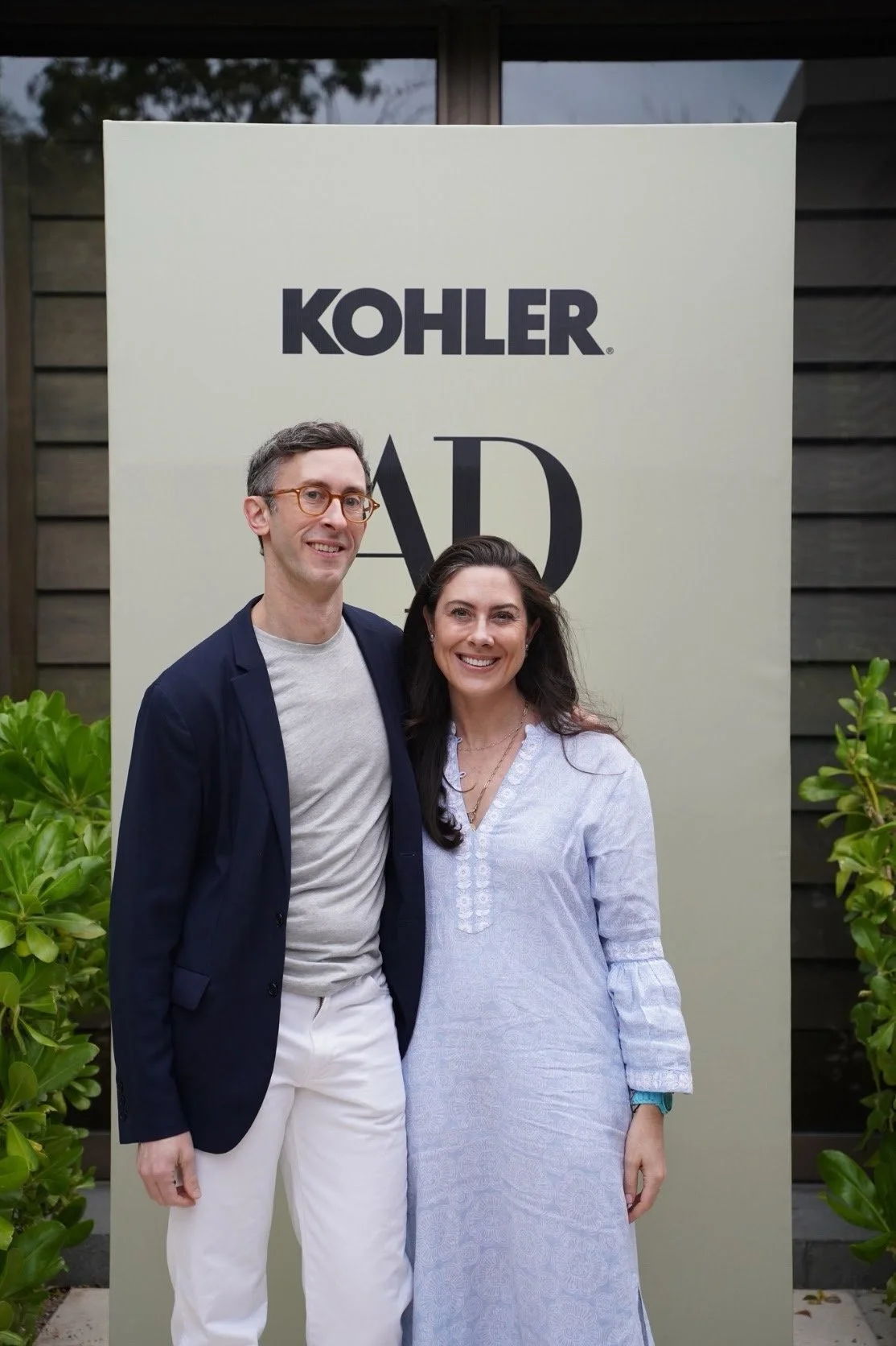 Two people smiling, standing in front of a sign that reads 'Kohler Award.' The man on the left wears glasses, a navy blazer, a light gray shirt, and white pants. The woman on the right has long dark hair, wearing a light blue dress. There are green p