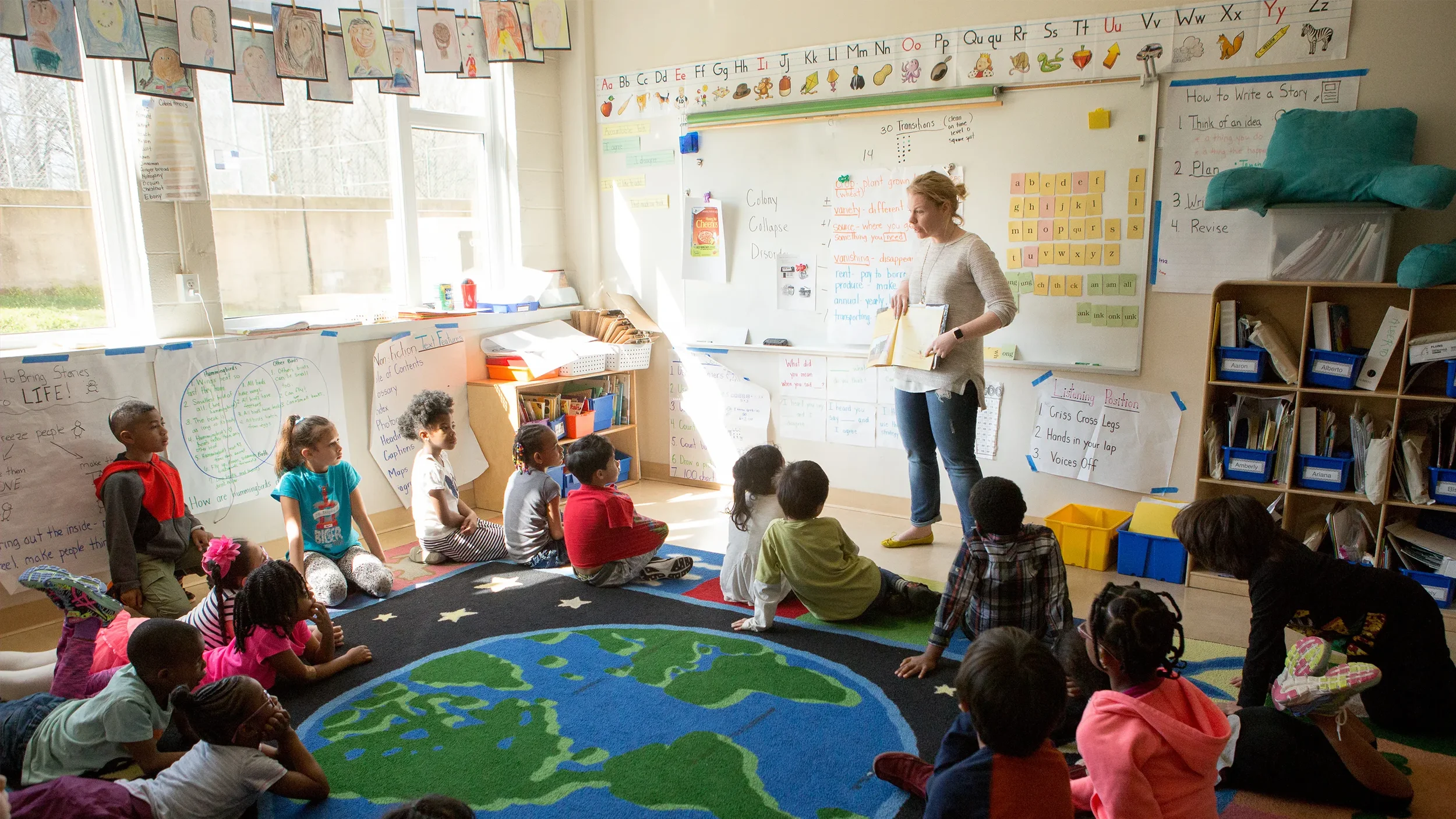 A teach at the front of the room and a classroom of young school children sitting on the floor watching her.