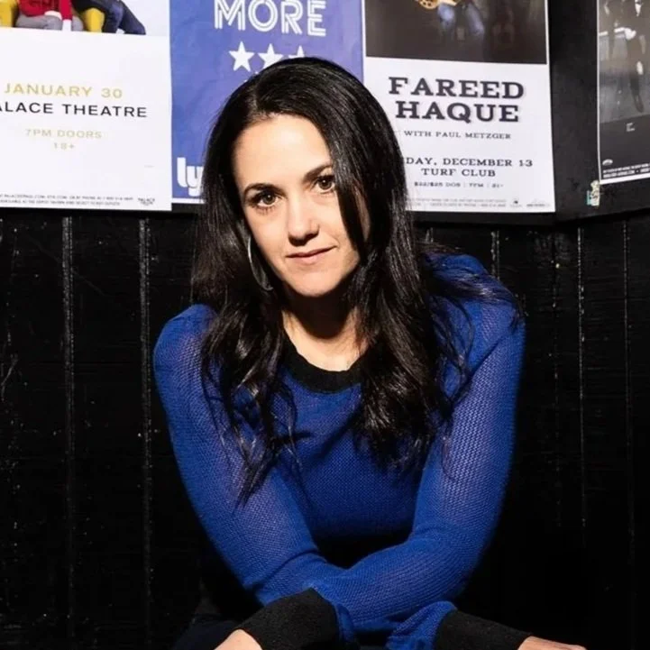 Woman 'DAYNA FRANK' with long dark hair in a blue sweater sitting against a black wall with concert posters behind her.