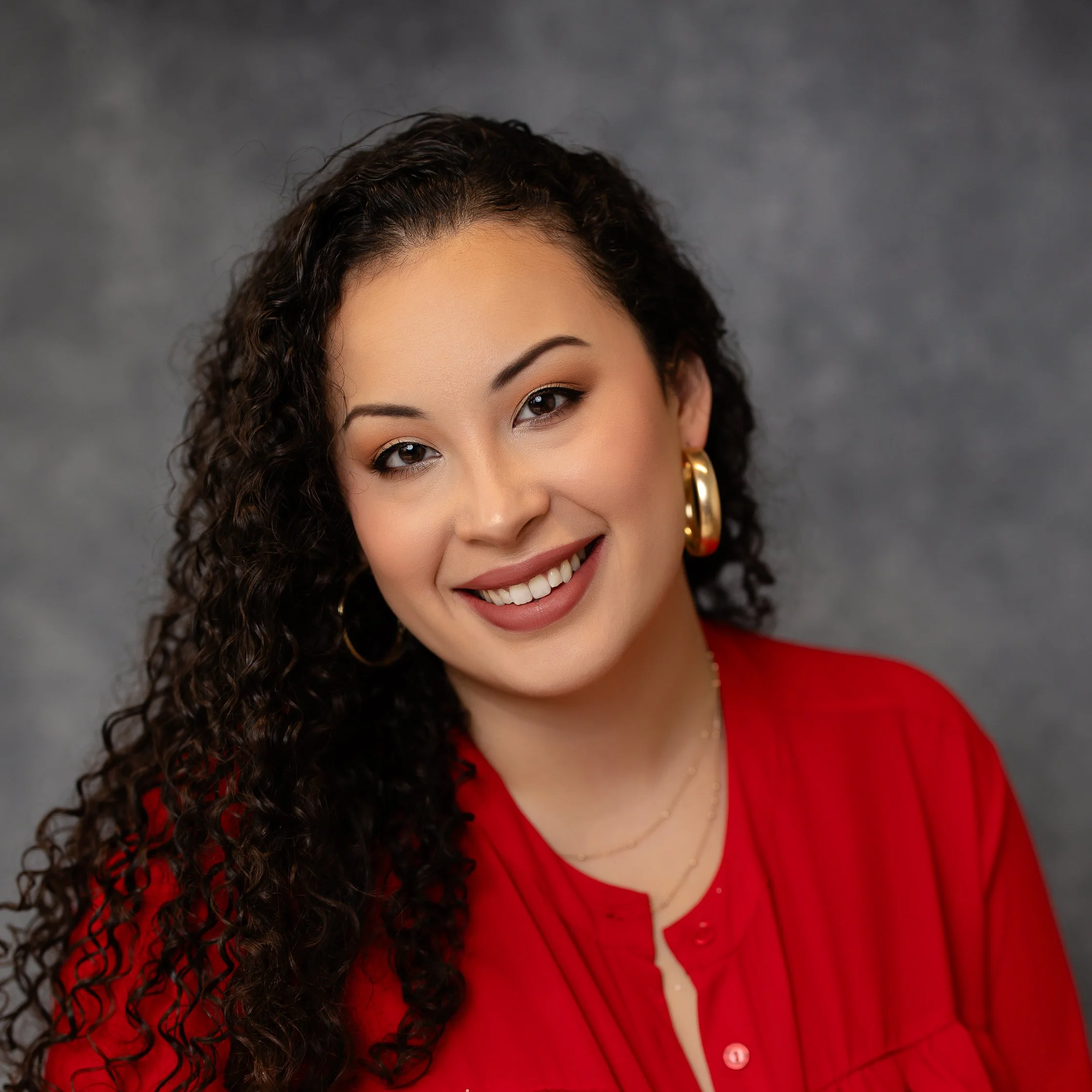 Woman 'LANI ECLATT' with curly dark hair, wearing gold hoop earrings and a red blouse, against a gray background.