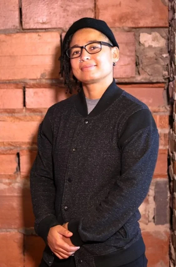 Woman 'TOBI PARKS' with glasses, short dreadlocks, wearing a black beanie and a black jacket, standing against a brick wall.
