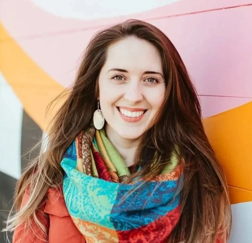 Woman 'DIANA WEISHAAR' with long brown hair standing in front of a colorful painted wall, wearing a multicolored scarf and a reddish shirt.