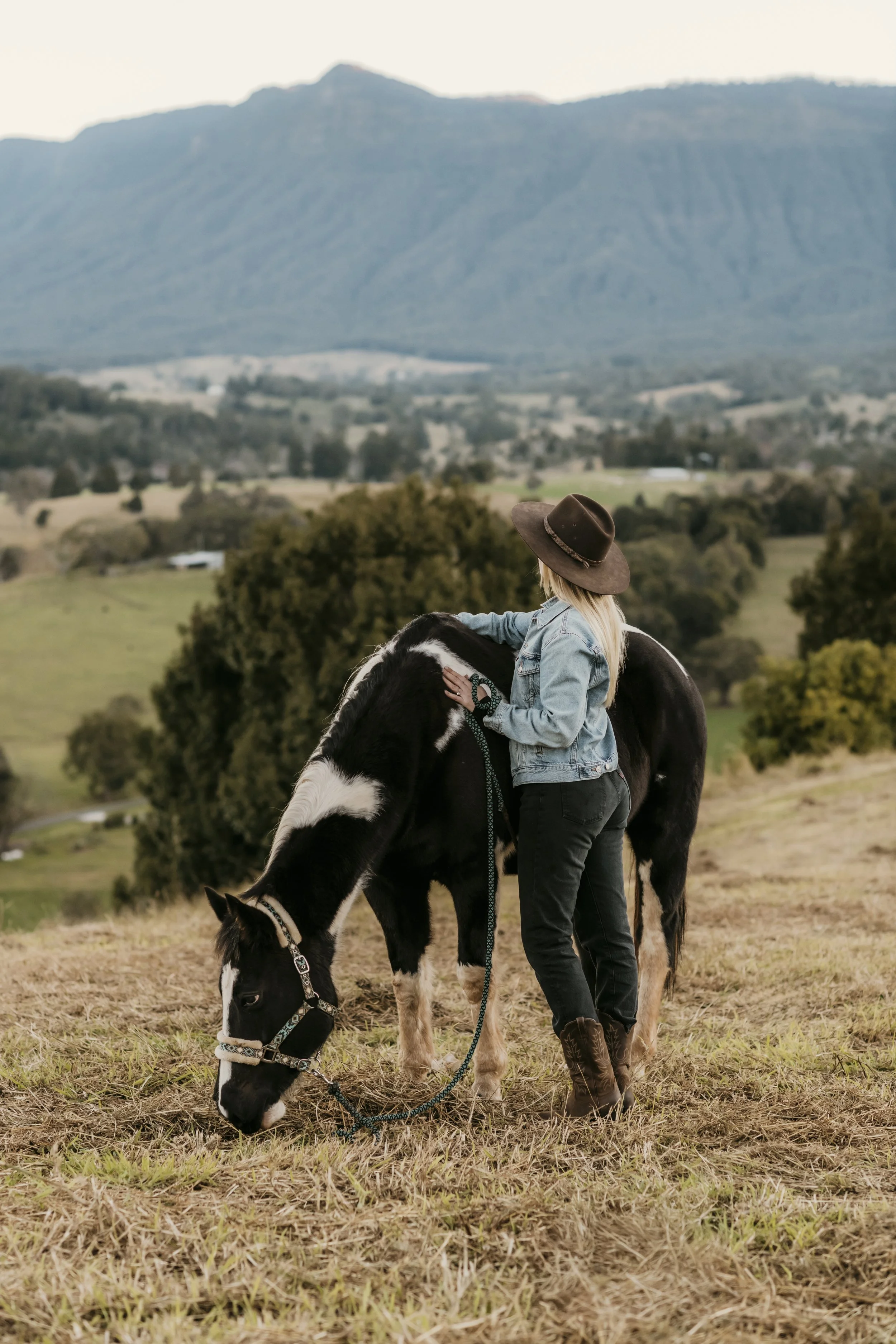 A person with blonde hair wearing a wide-brimmed hat, denim jacket, and dark jeans is standing outdoors in a pasture, petting a black and white horse that is grazing with its head lowered, in front of a scenic landscape with rolling hills and mountains.
