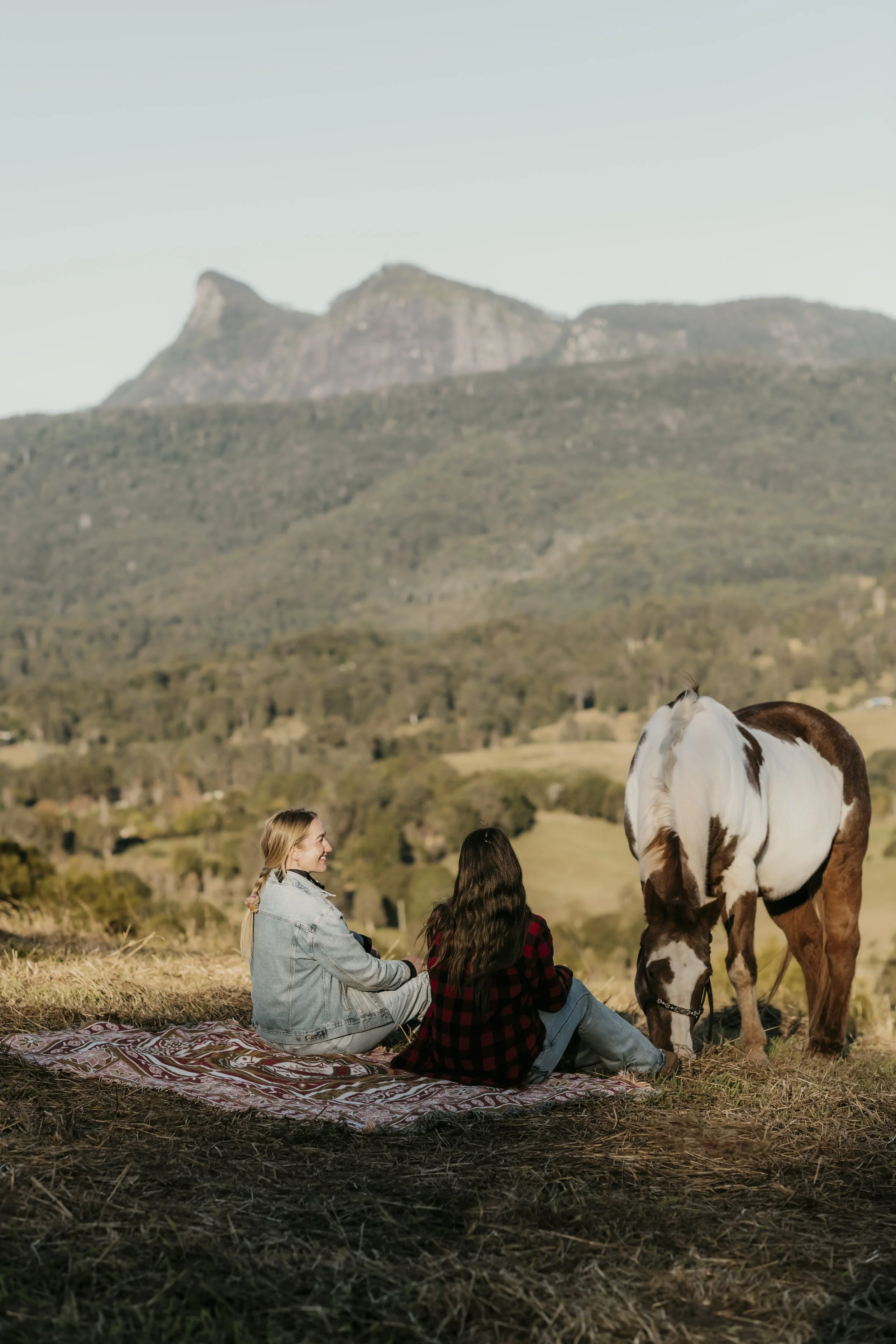 Two women sitting on a blanket in a grassy field, with a white and brown horse nearby, in a rural landscape with hills and mountains in the background.