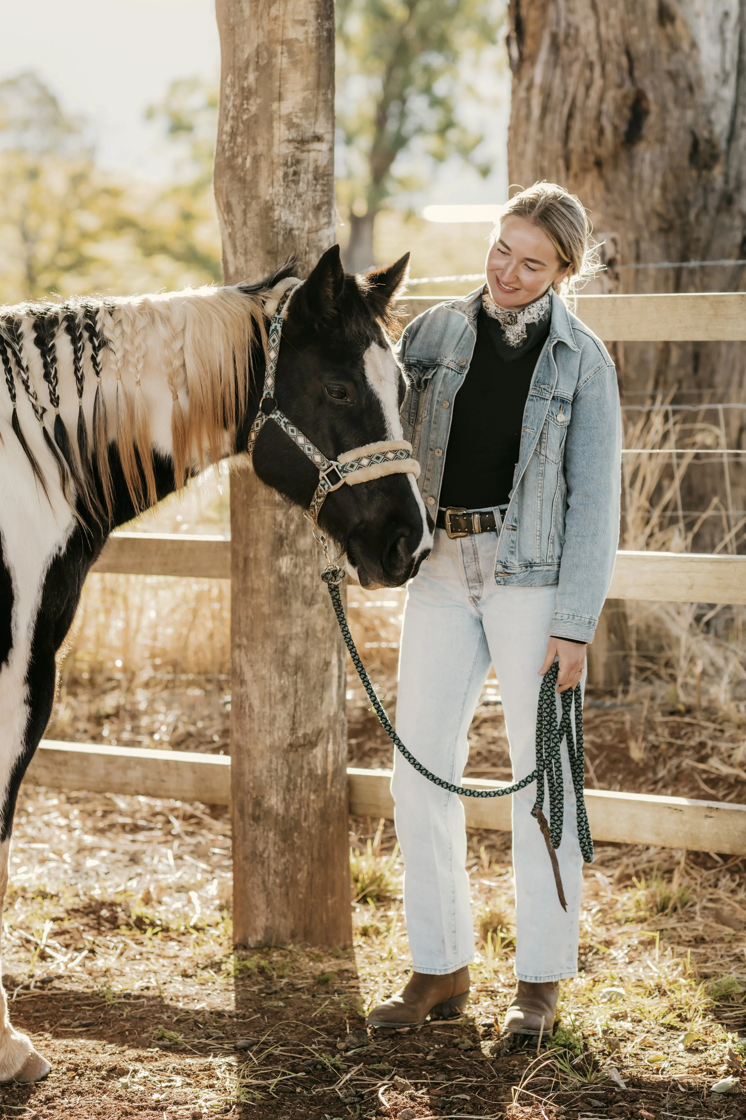A young woman in a denim jacket and light blue jeans smiling at a black and white horse in a fenced outdoor area with trees and sunlight.