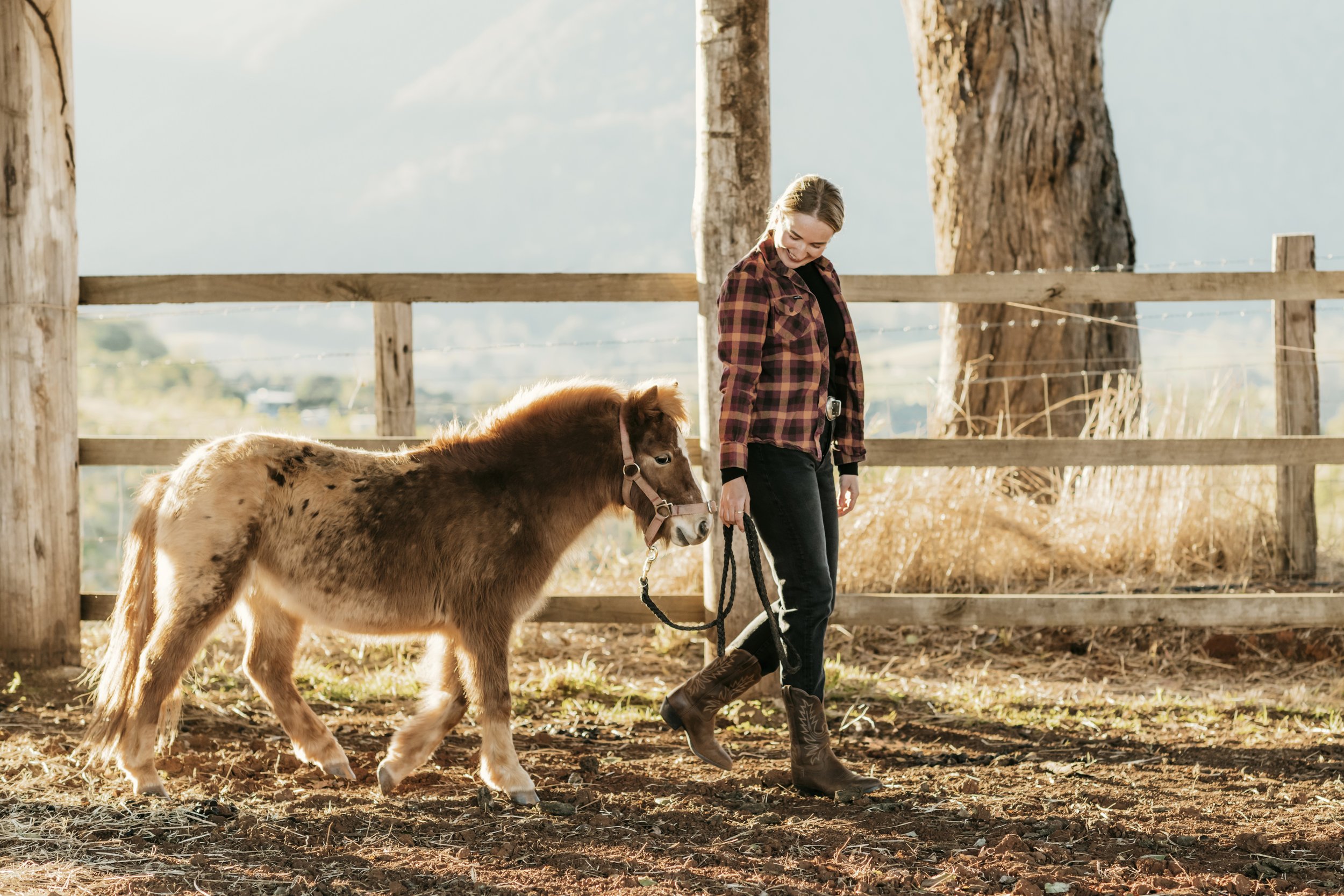 A young woman with blonde hair smiling and walking a small brown and white pony in a wooden-fenced outdoor area during the daytime. The background includes tall trees and a scenic landscape with mountains.