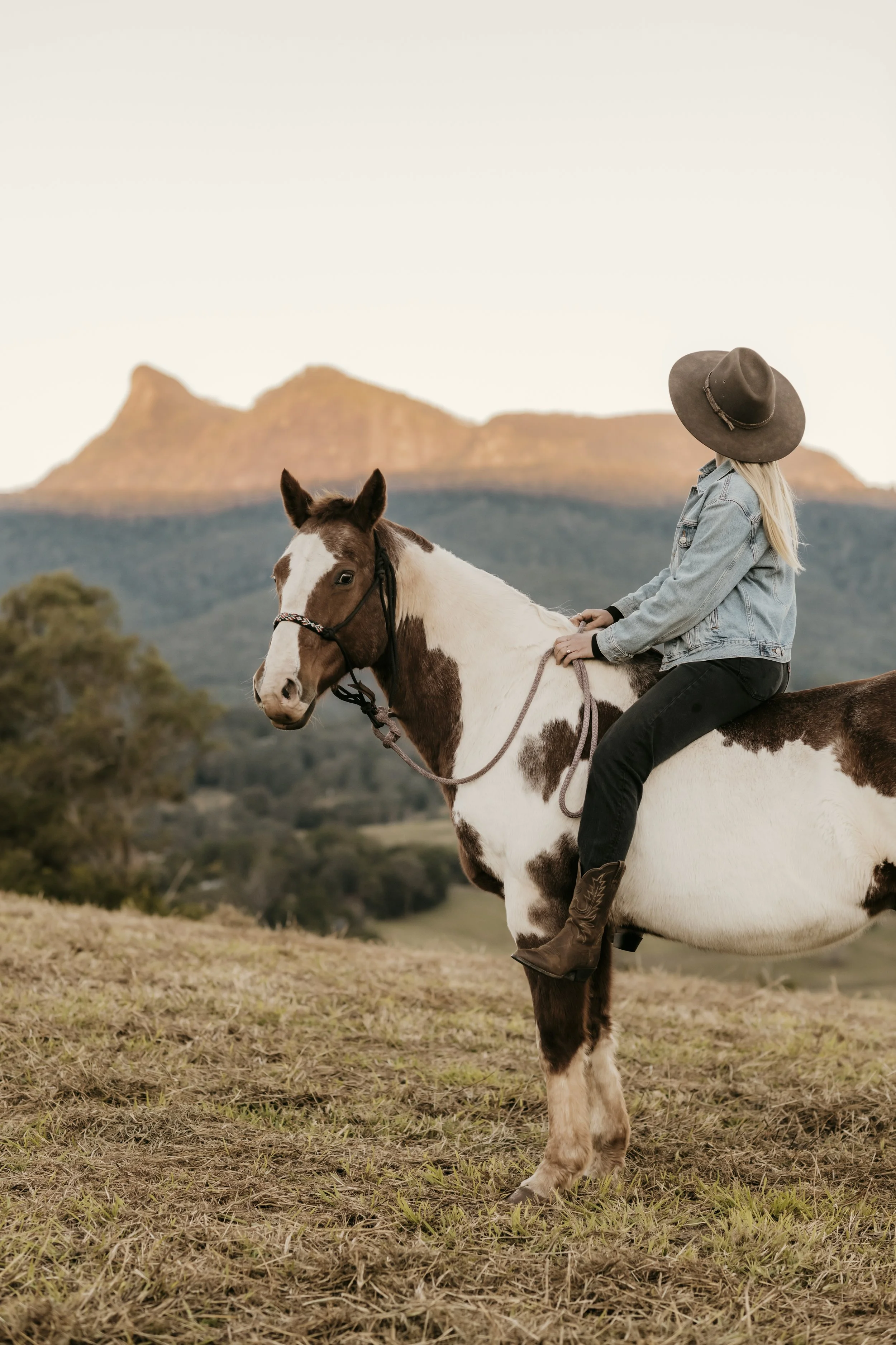 A woman wearing a large hat, denim jacket, and black pants rides a white and brown pinto horse in an open field with mountains in the background at sunset.
