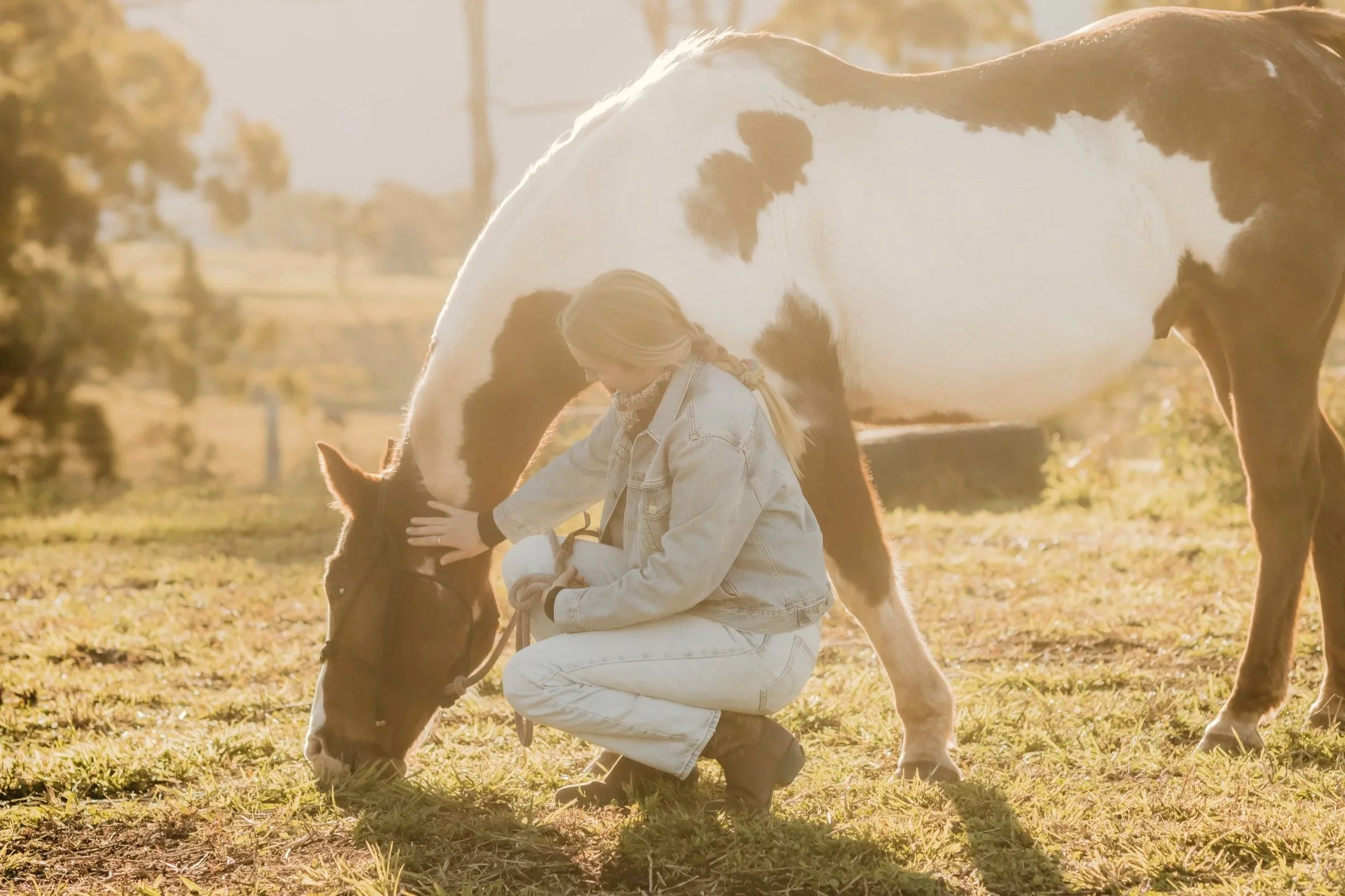 A woman with blonde hair tied in braids, wearing a denim jacket and white pants, kneeling on the grass while caring for two small ponies in a sunlit outdoor setting with trees in the background.