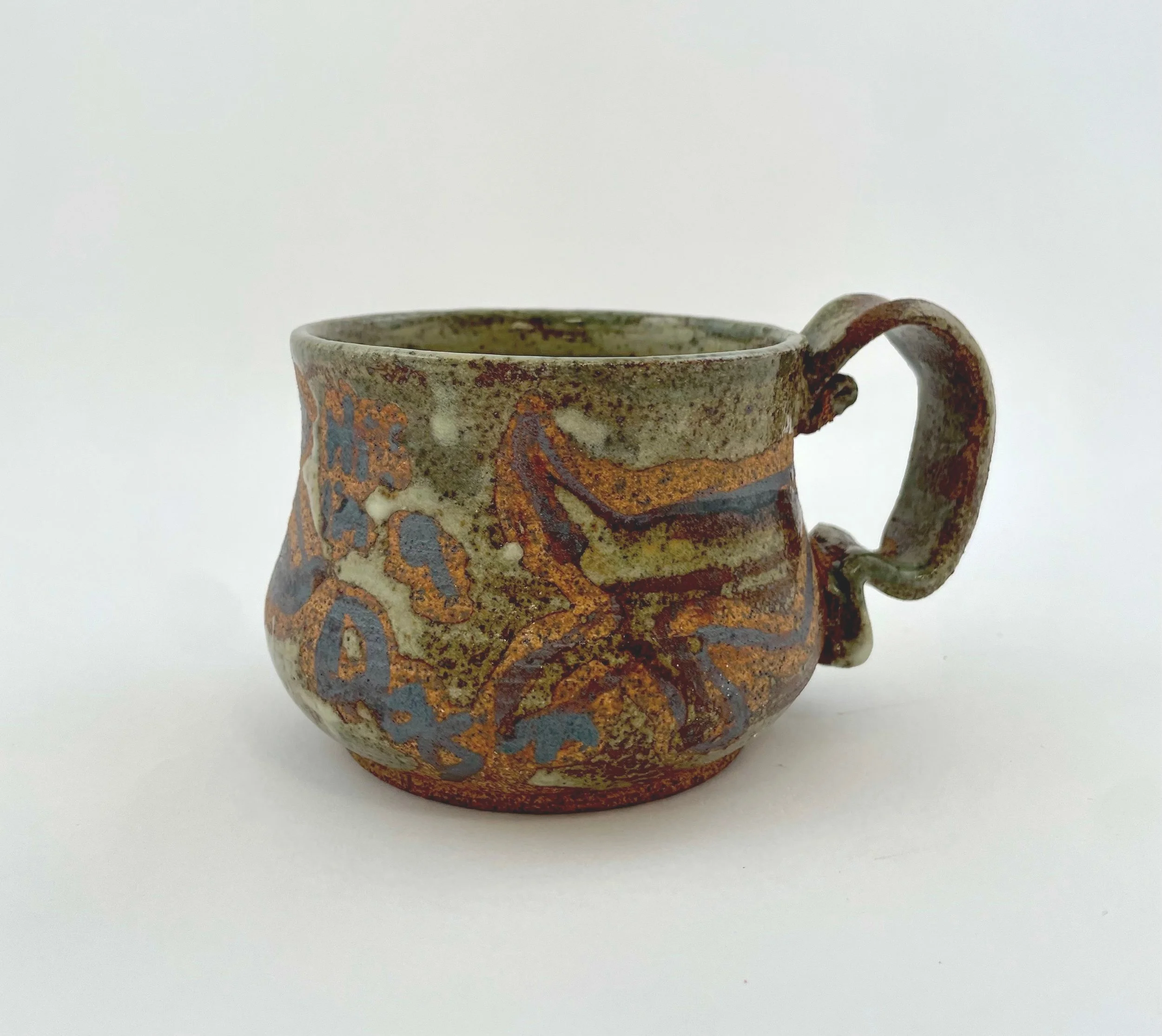 A small ceramic mug with a textured, earthy design and abstract patterns in shades of blue, orange, and brown. The mug has a rounded shape and a curved handle.