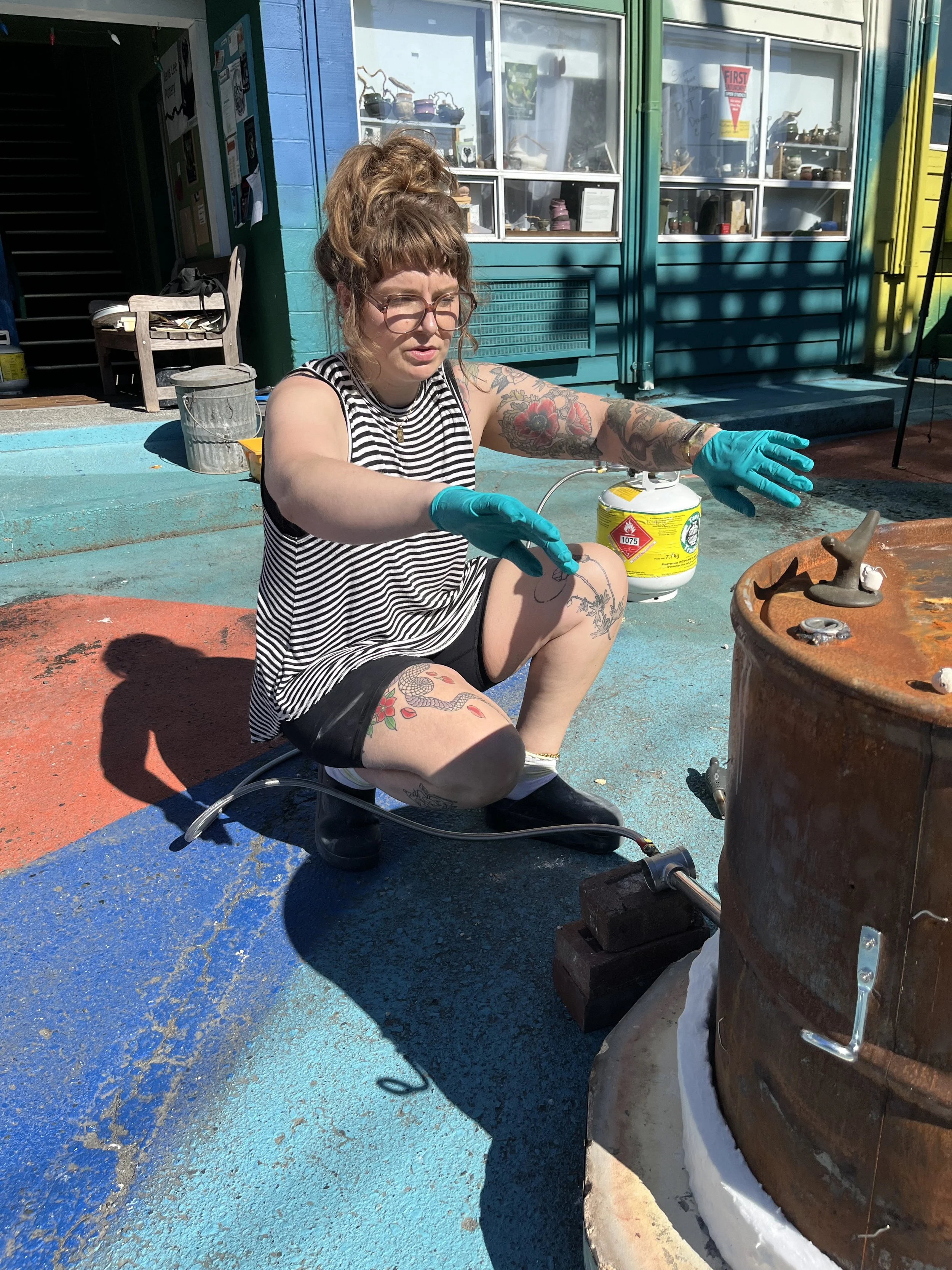 A woman with tattoos, wearing glasses, a striped sleeveless top, black shorts, and blue gloves, crouches outdoors near a rusty metal barrel, holding a tool and working with a gas canister and equipment in front of a colorful building with blue, green, and yellow walls.