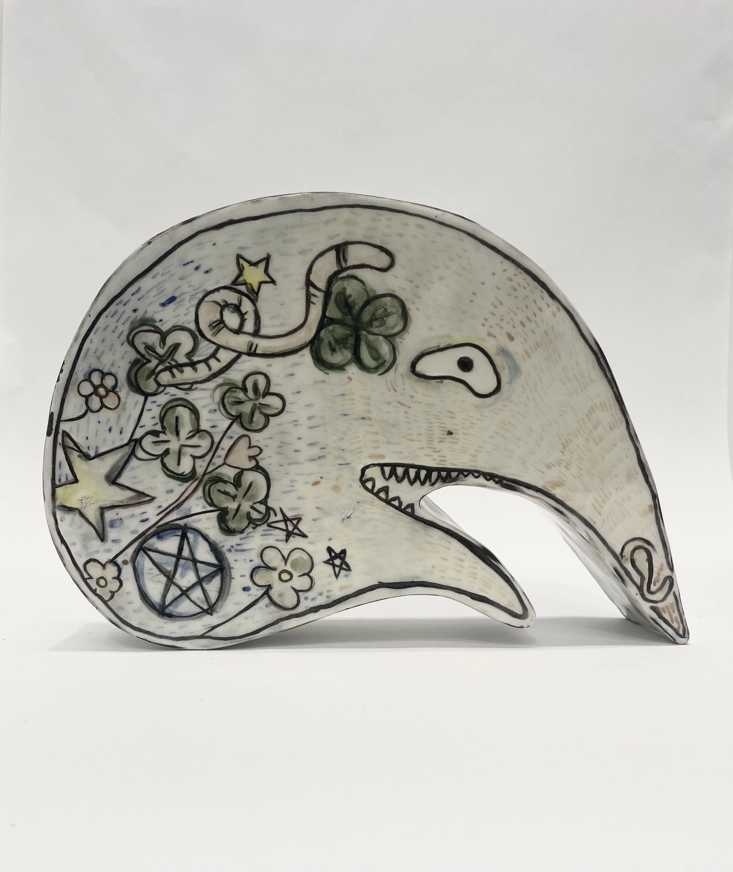A ceramic crescent-shaped dish decorated with whimsical drawings of a snake, flowers, a star, a pentagram, and a small creature, all outlined in black with some colored accents.