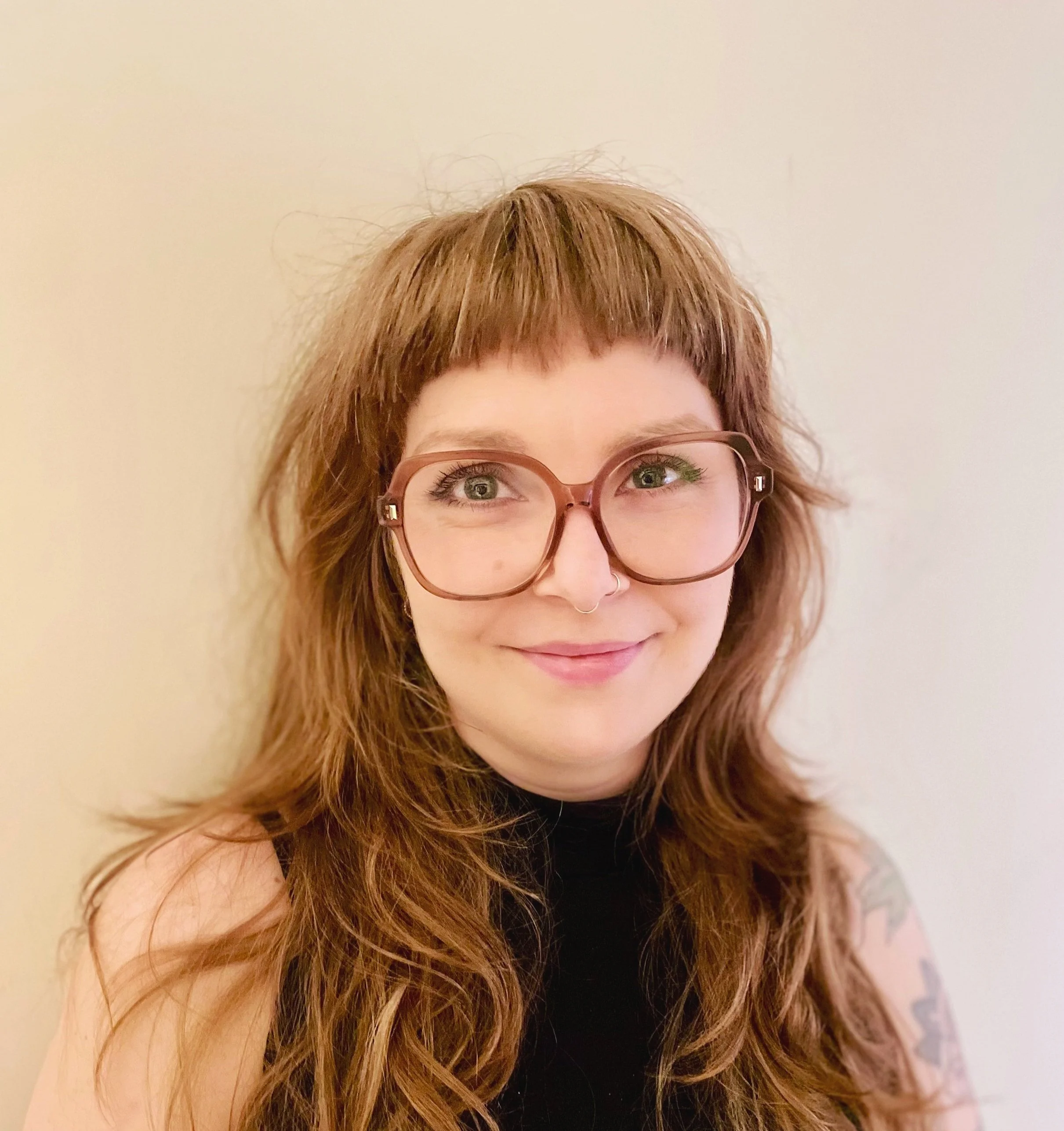A woman with red hair, glasses, and a septum piercing smiling against a plain wall.