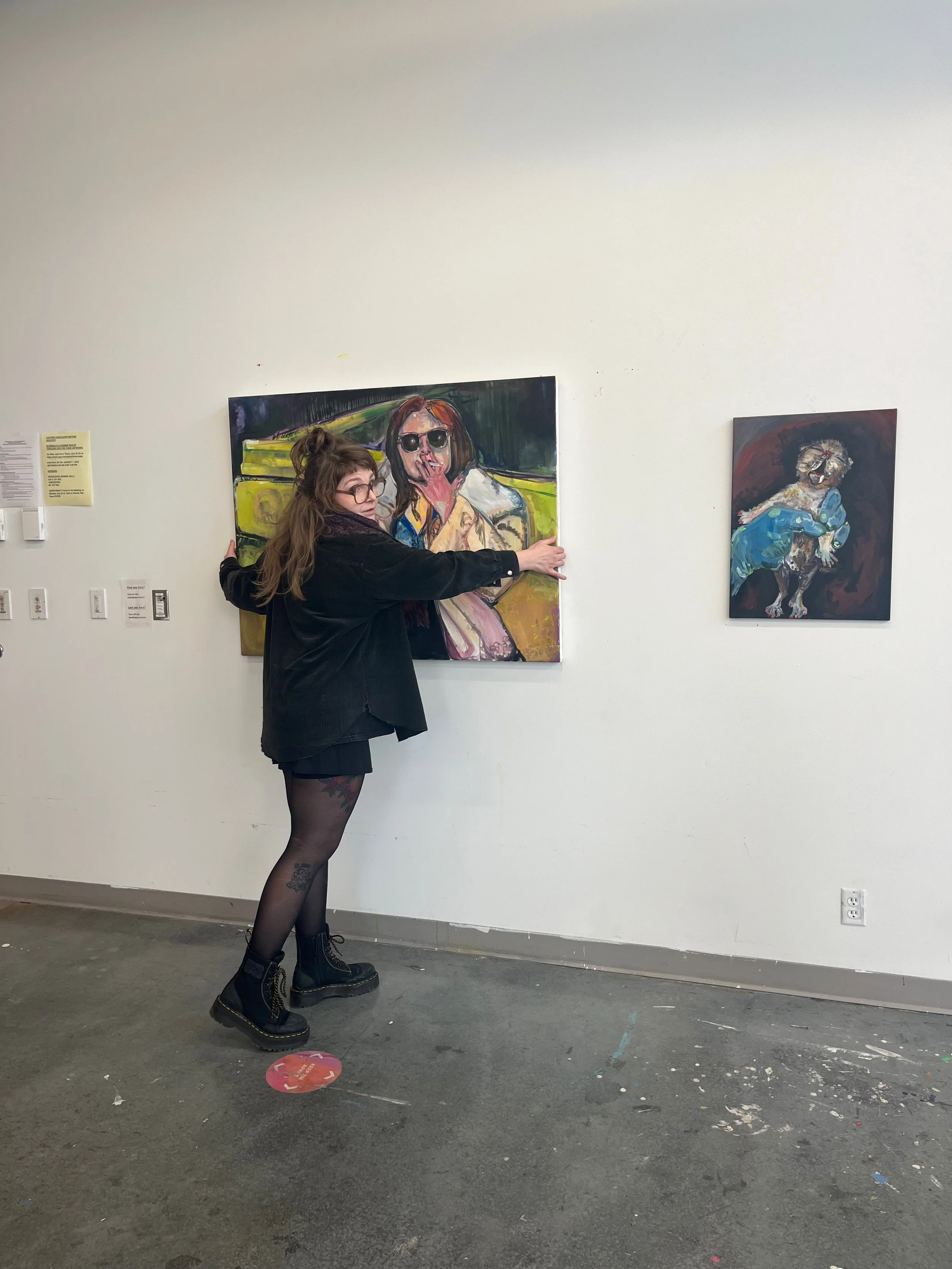 A woman with long brown hair and glasses is standing in front of a white gallery wall, touching a colorful portrait painting of a woman with sunglasses. She is wearing a black jacket, black skirt, patterned tights, and black boots. There is another smaller painting of a woman on the wall to her right.