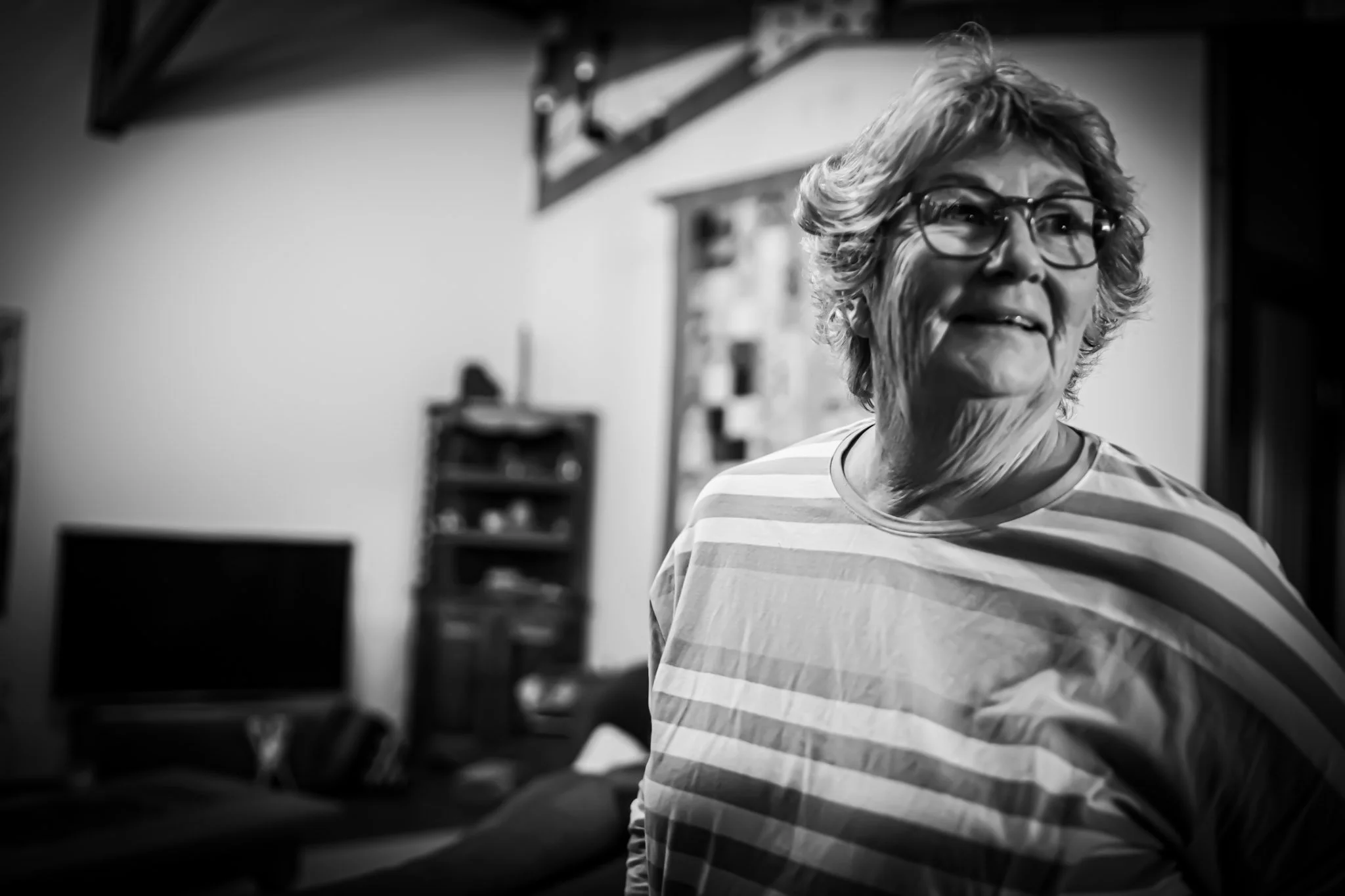 A smiling elderly woman with glasses and curly hair is wearing a striped shirt and looking to her right in a living room.