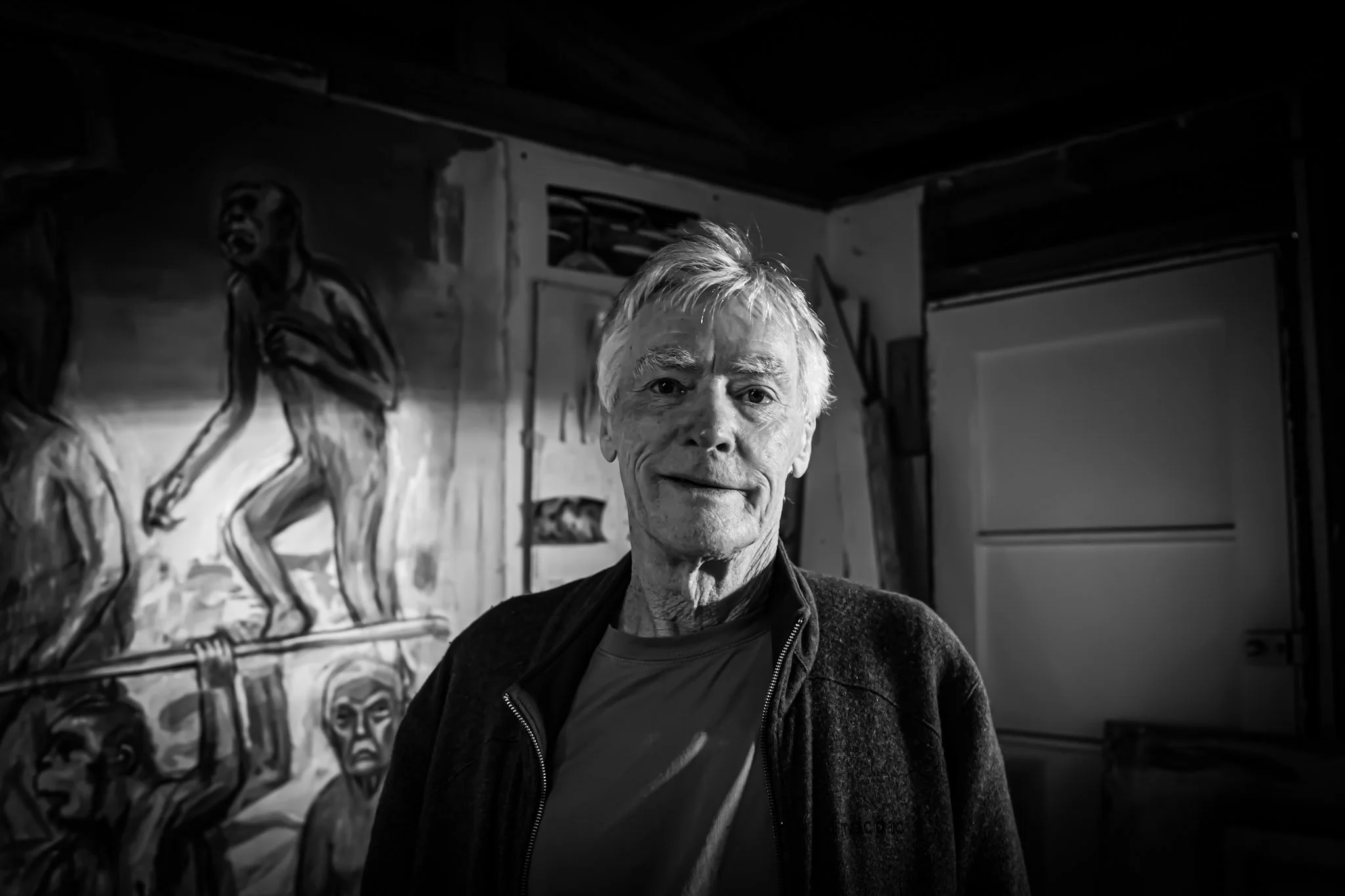Black and white photo of an elderly man with light-colored hair standing in front of a painting of a dancer.