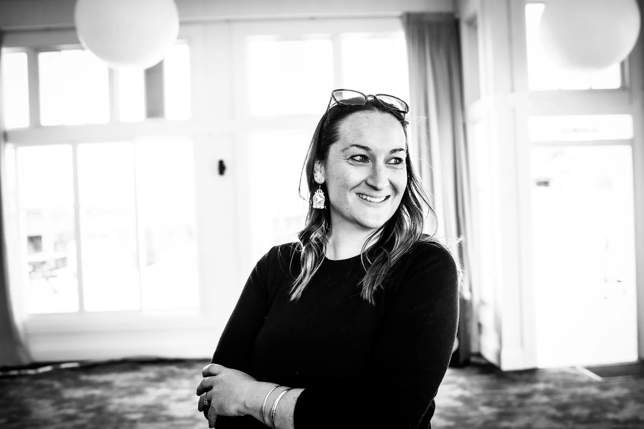 Black and white photo of a woman smiling, wearing earrings, a ring, and glasses on her head, standing indoors near large windows.