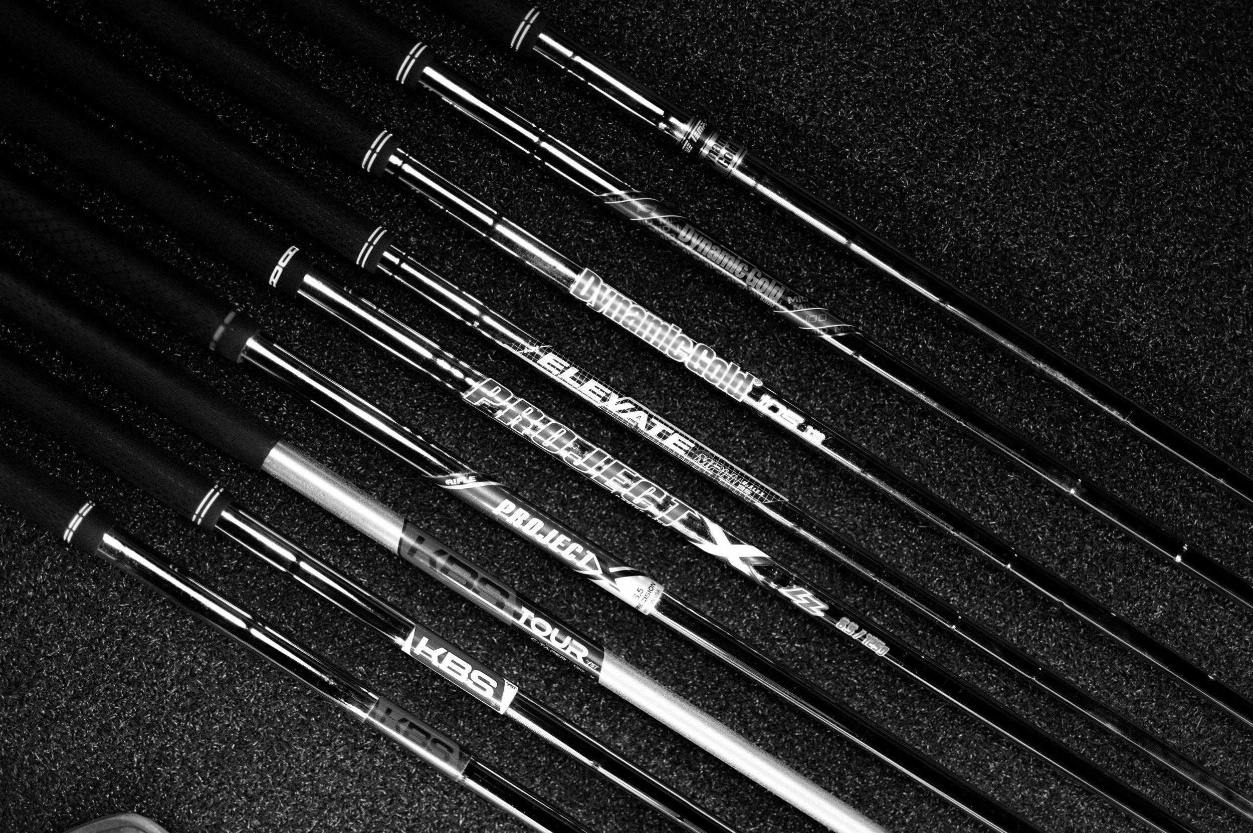 Six golf clubs with black grips laid out on a textured black surface, arranged diagonally.