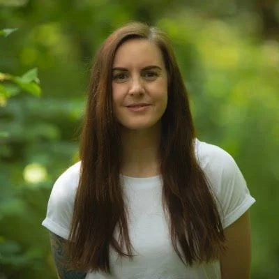 Young woman with long brown hair wearing a white t-shirt outdoors in a green, lush environment.