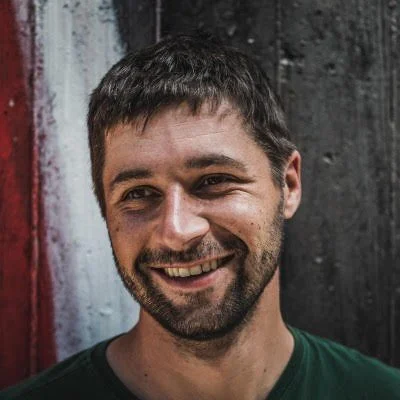 A smiling man with short dark hair, a beard, and wearing a green shirt standing in front of a background with dark and white vertical stripes.