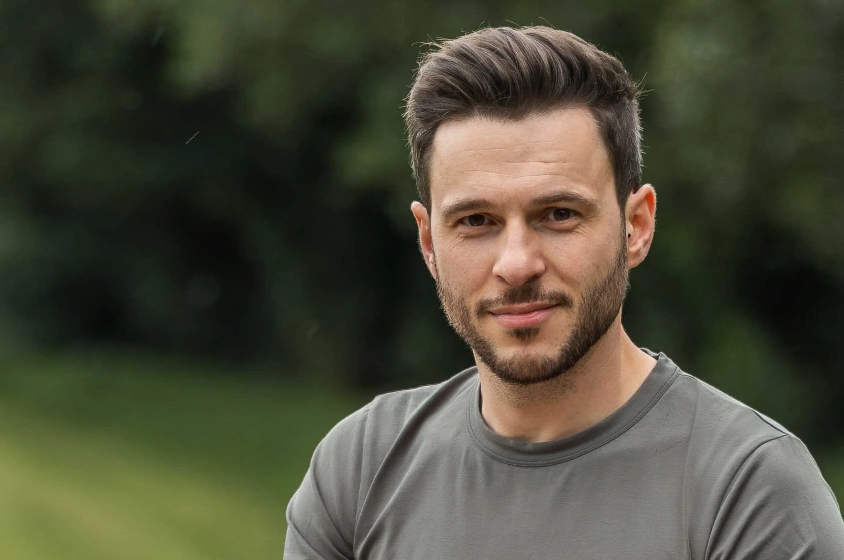 A young man with short dark hair, beard, and a mustache, wearing a gray t-shirt, outdoors with green blurred background.