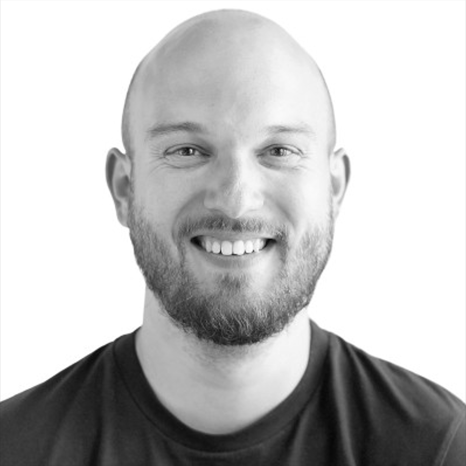 A smiling man with a beard, wearing a black shirt, looking directly at the camera, against a plain background.